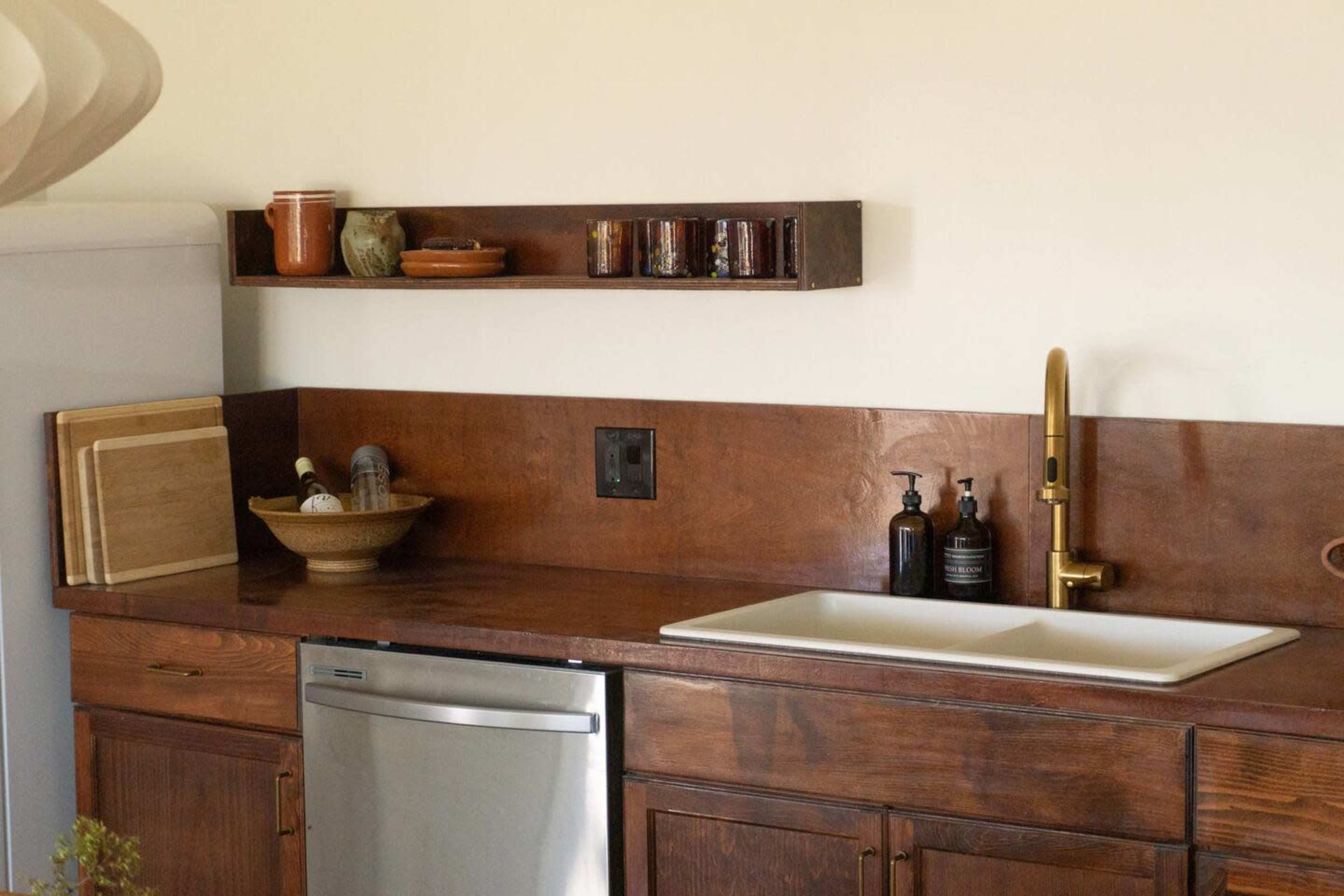 The image shows a kitchen countertop with a stainless steel dishwasher, a ceramic sink, wooden shelves holding jars, and various kitchen items neatly arranged.