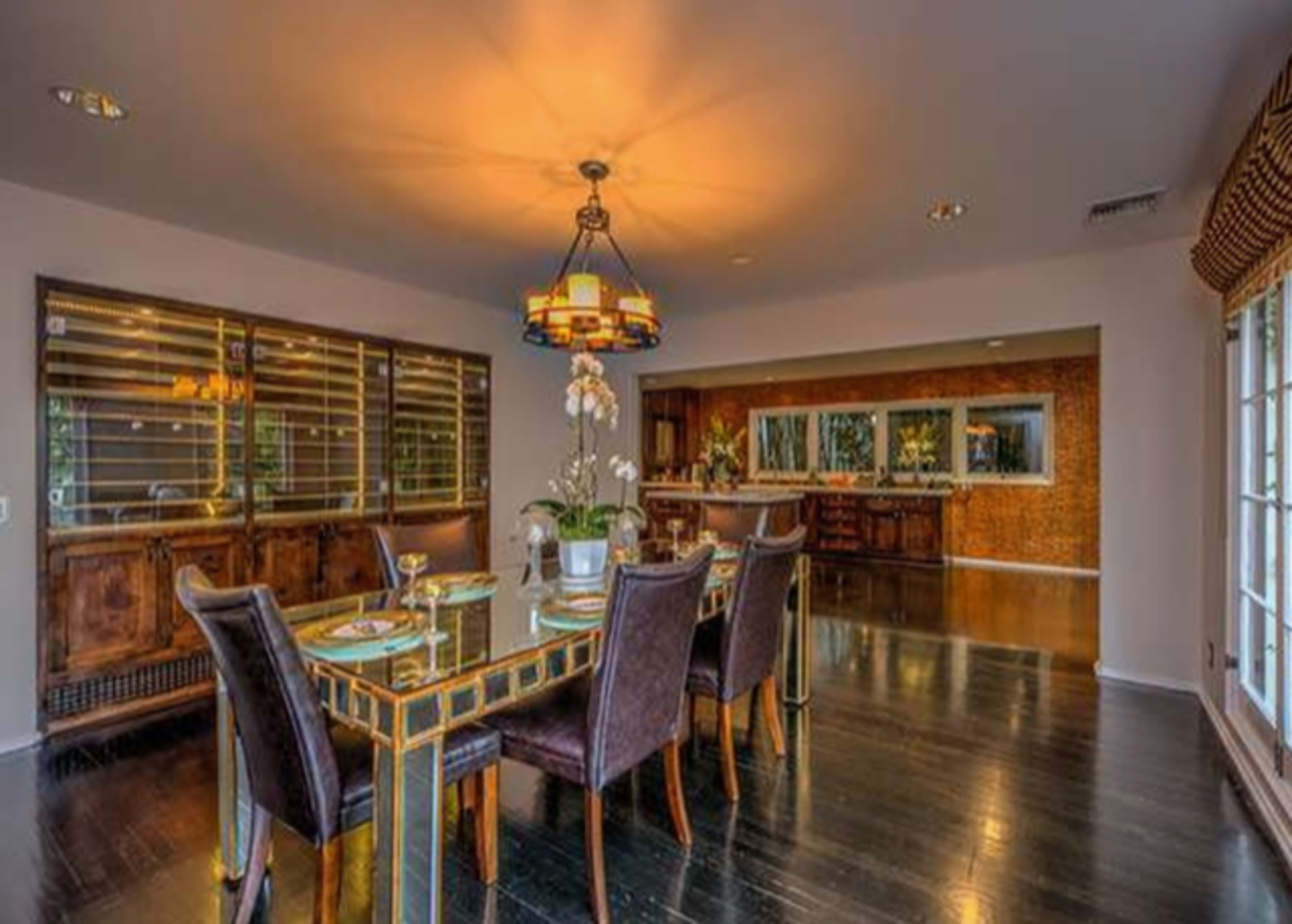 The image shows a dining room with a long table set for a meal, surrounded by dark upholstered chairs, and features a wooden buffet and an open kitchen area in the background.
