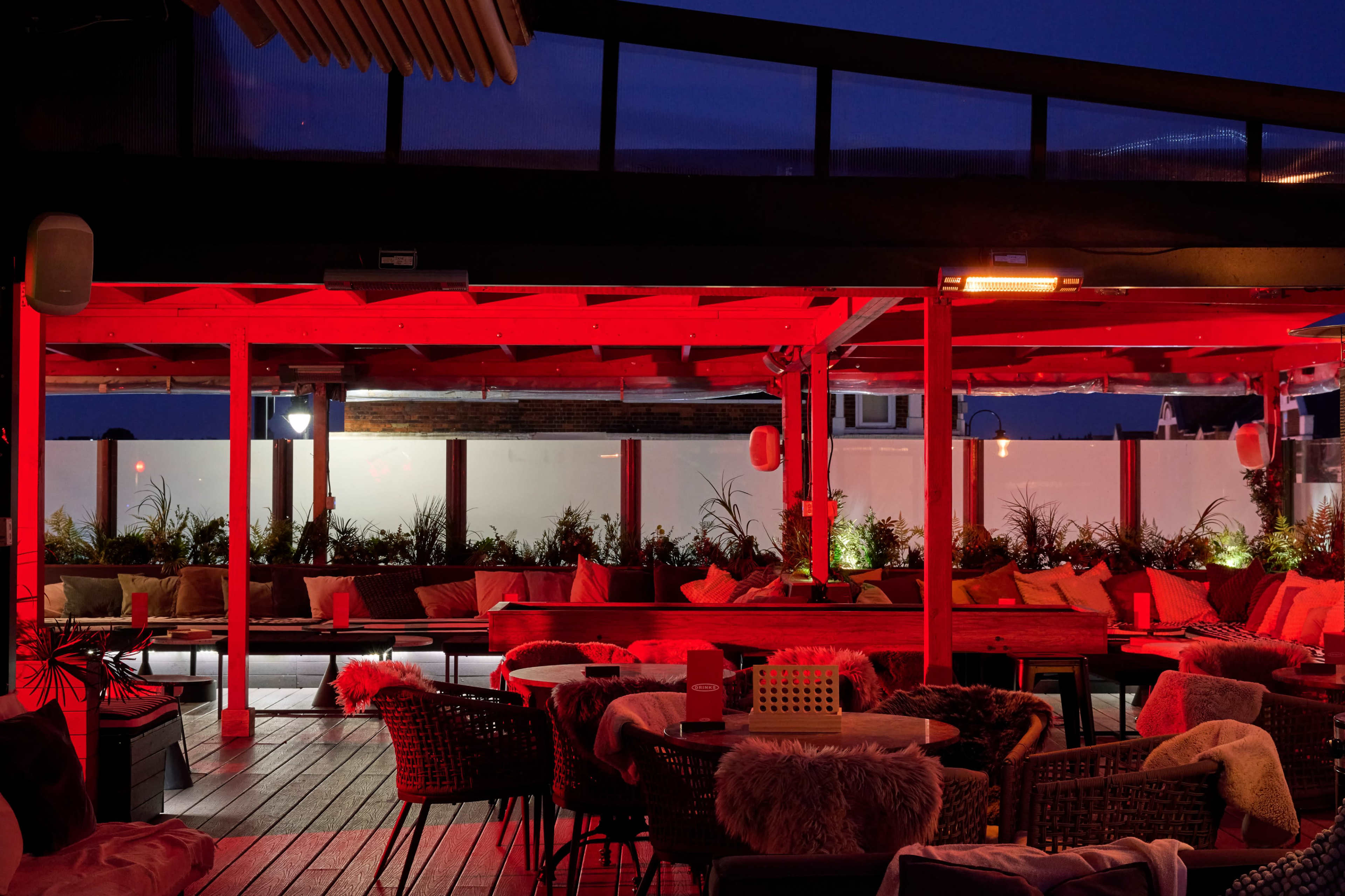 The image shows a rooftop lounge area with red lighting, featuring seating cushions and plants in decorative pots.