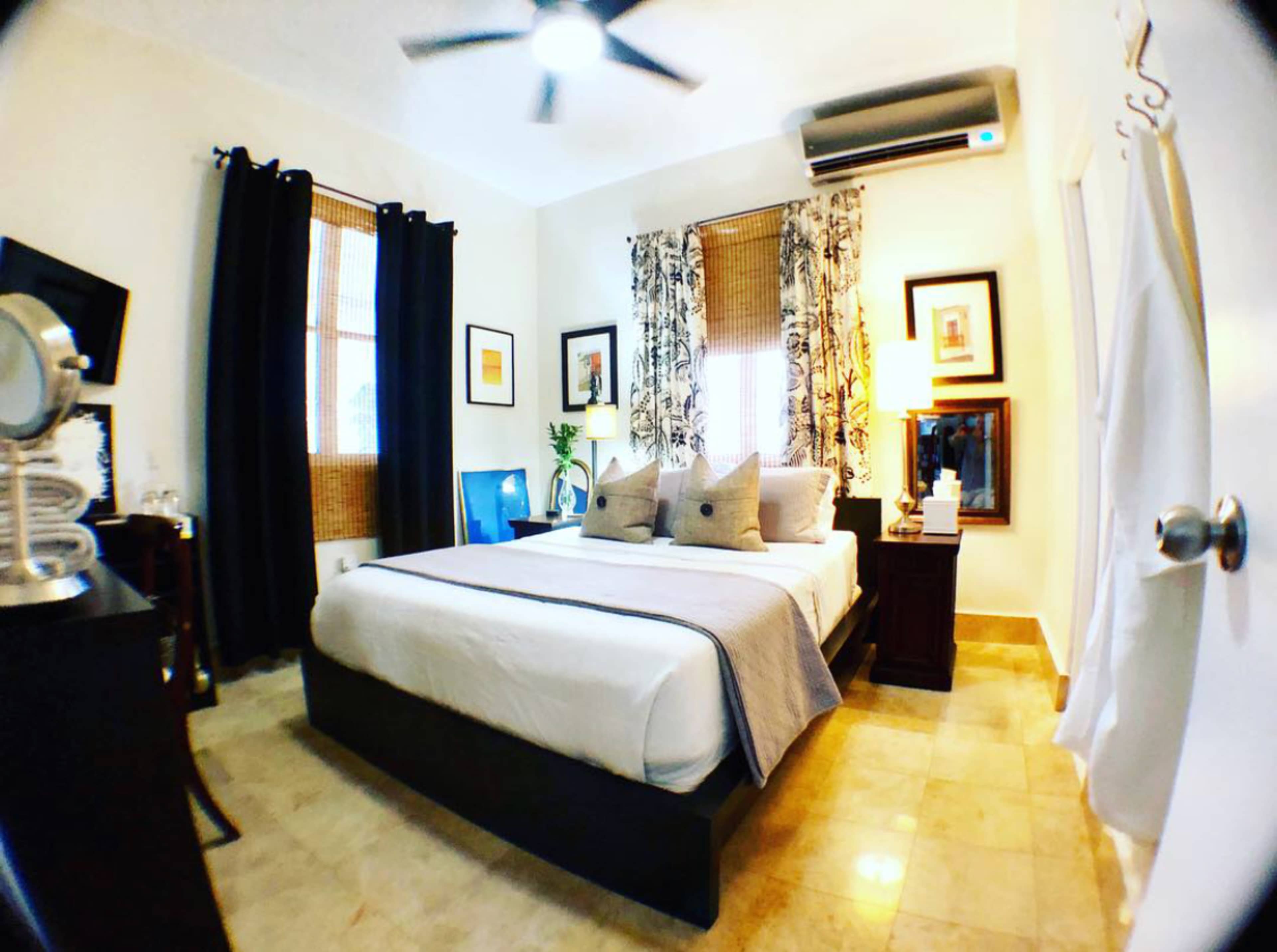 The image shows a well-decorated bedroom featuring a queen-sized bed, window treatments, and a small seating area with a blue chair.