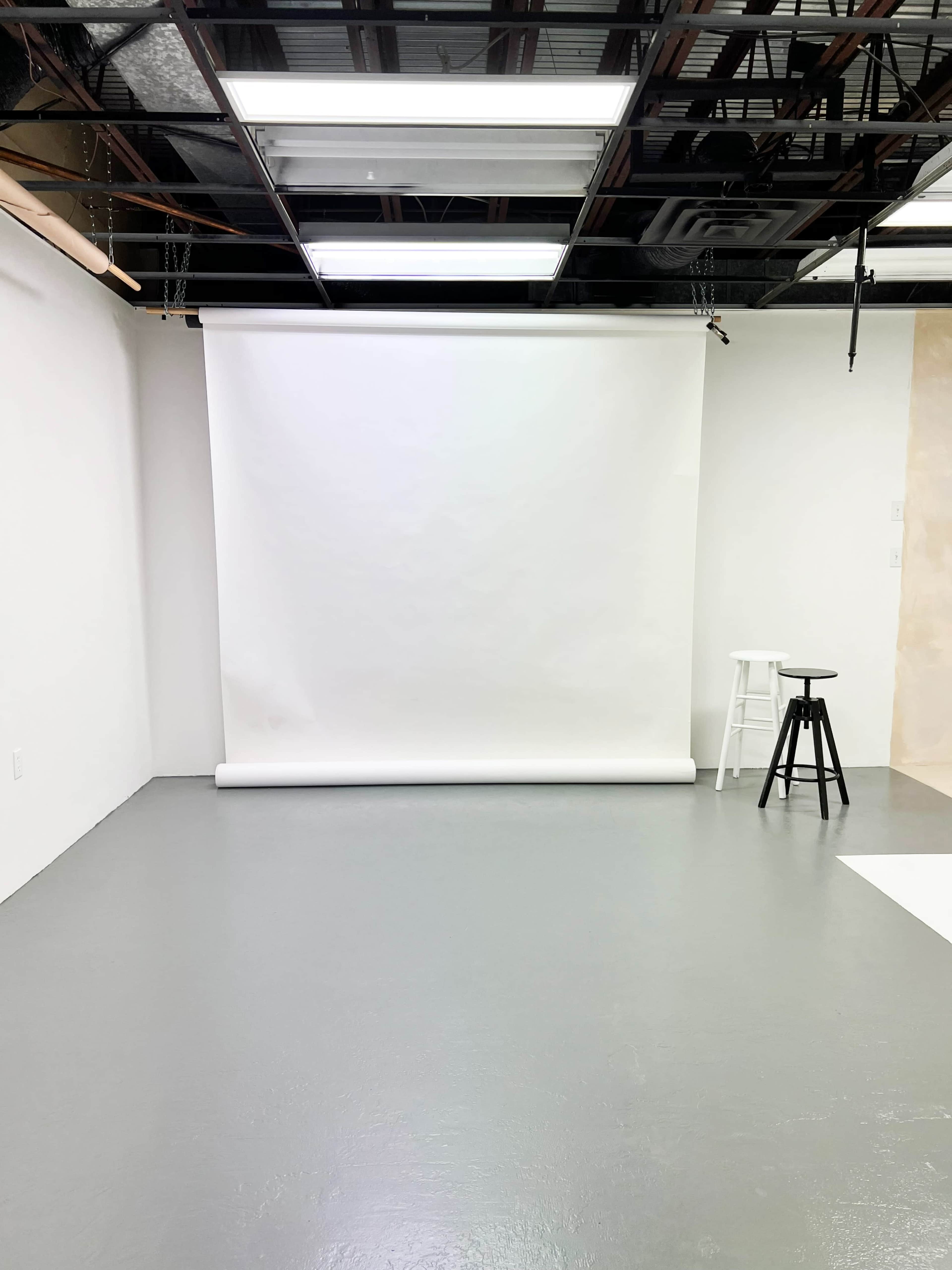 The image depicts a minimalistic photography studio with a white backdrop on a roll, a bare floor, and a single black stool.