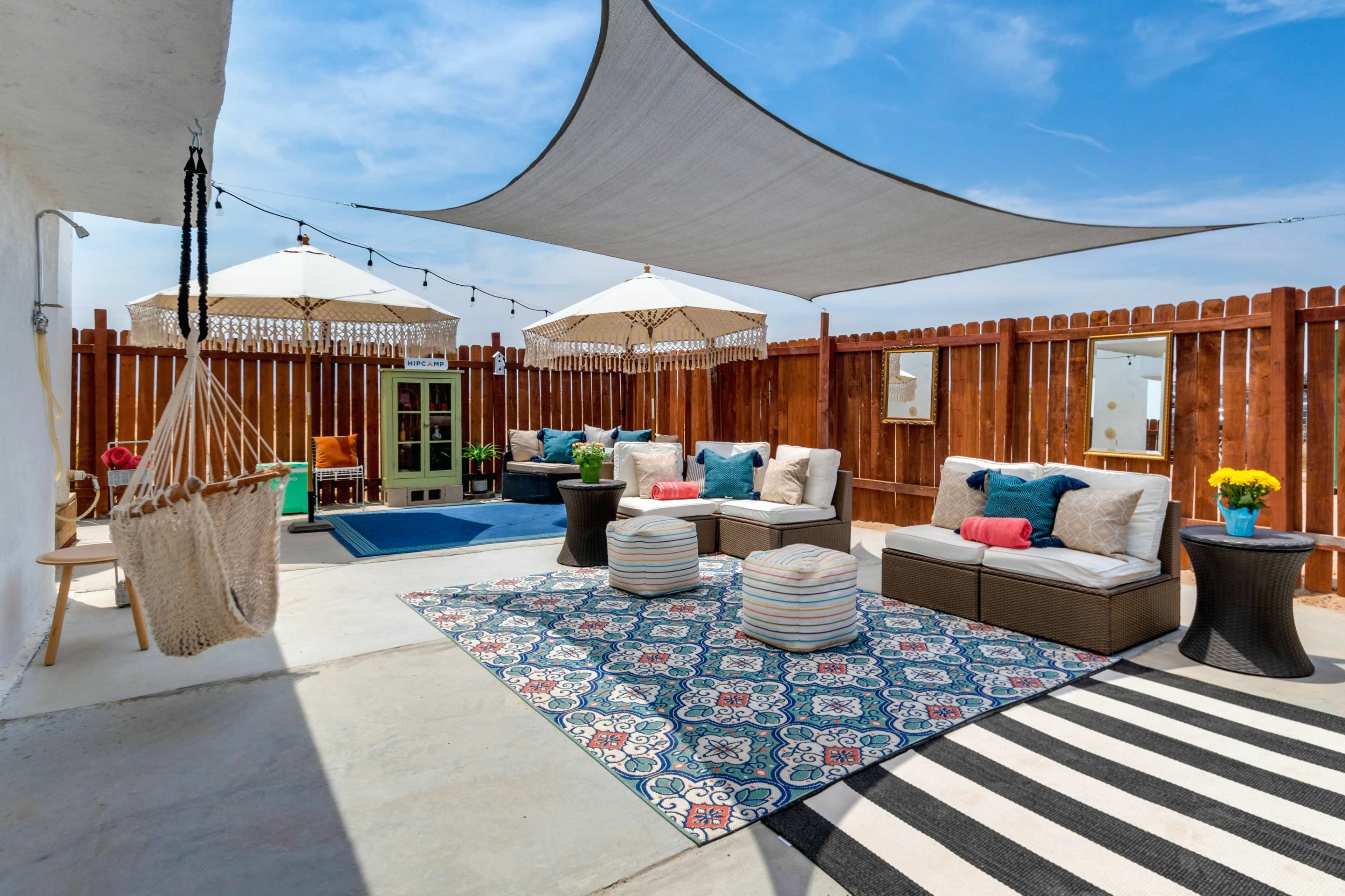 A patio area features a shaded seating arrangement with plush cushions, decorative umbrellas, and a patterned rug, surrounded by a wooden fence.