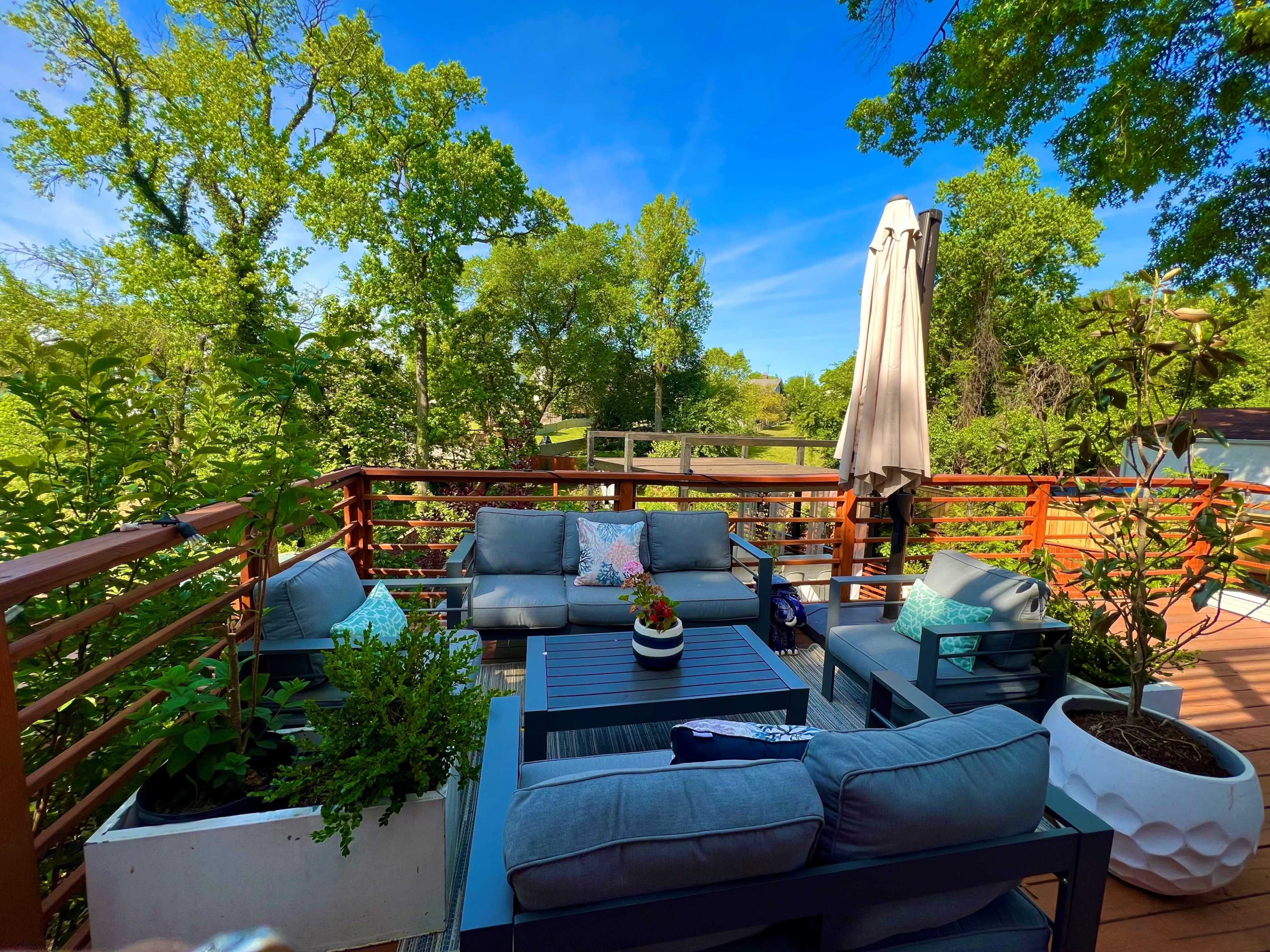 A comfortable outdoor seating area with couches and a large umbrella is surrounded by greenery and a wooden deck.
