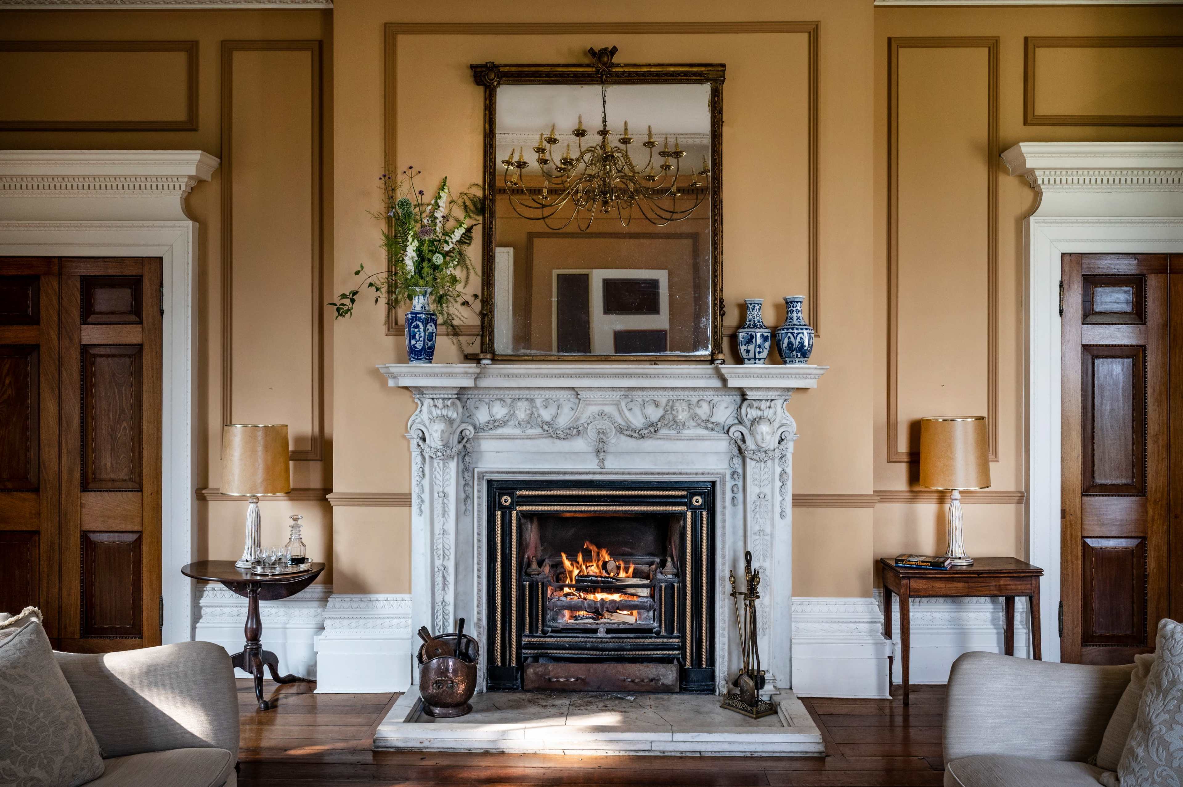 The image shows a fireplace with a chimneypiece, surrounded by elegant decor, including mirrors, vases, and wooden furniture.