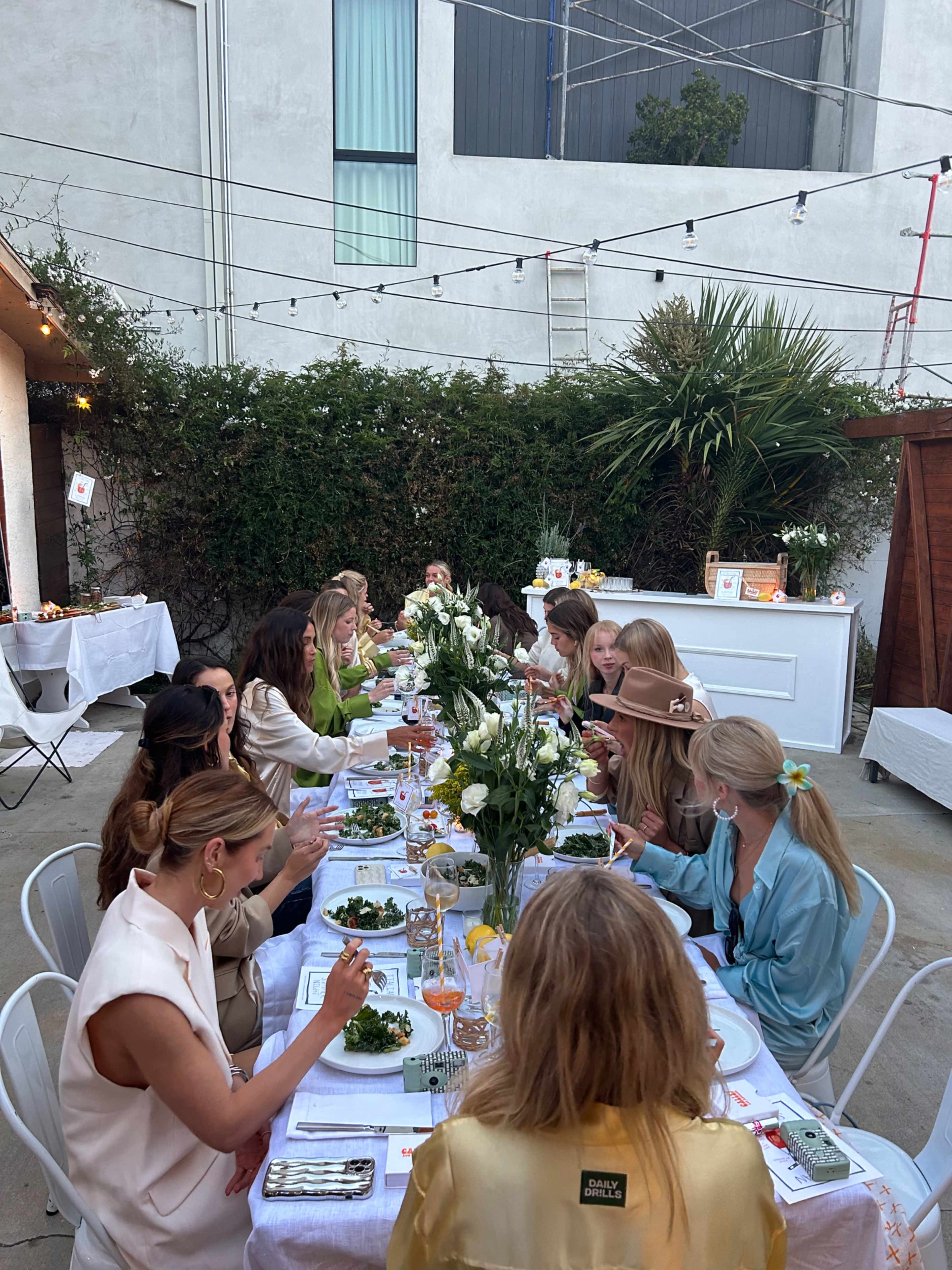 A long dining table is set outdoors with women dressed in various outfits, enjoying a meal amid greenery and string lights.