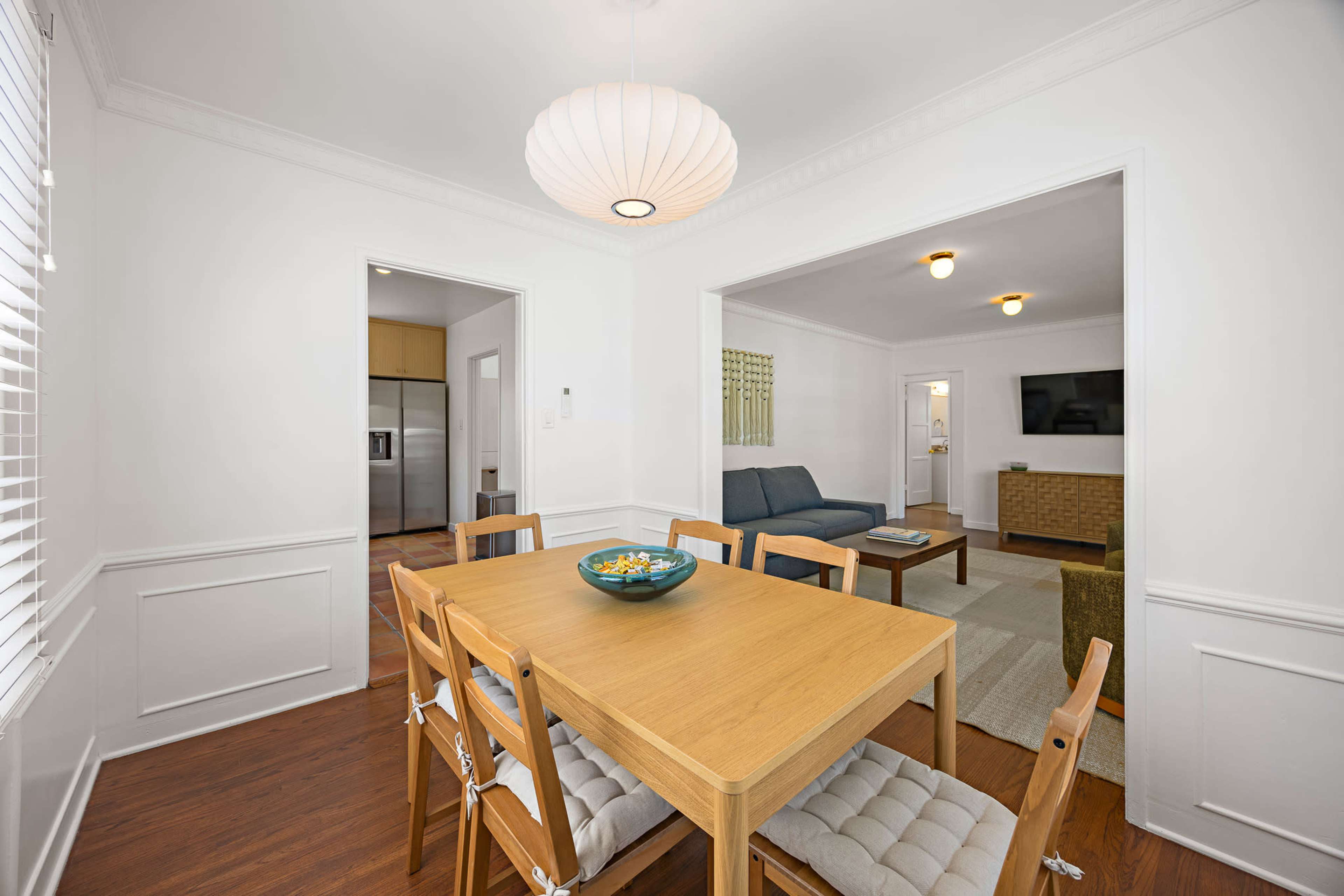 The image shows a well-lit dining area featuring a wooden table surrounded by chairs, with doorways leading to a kitchen and a living room.