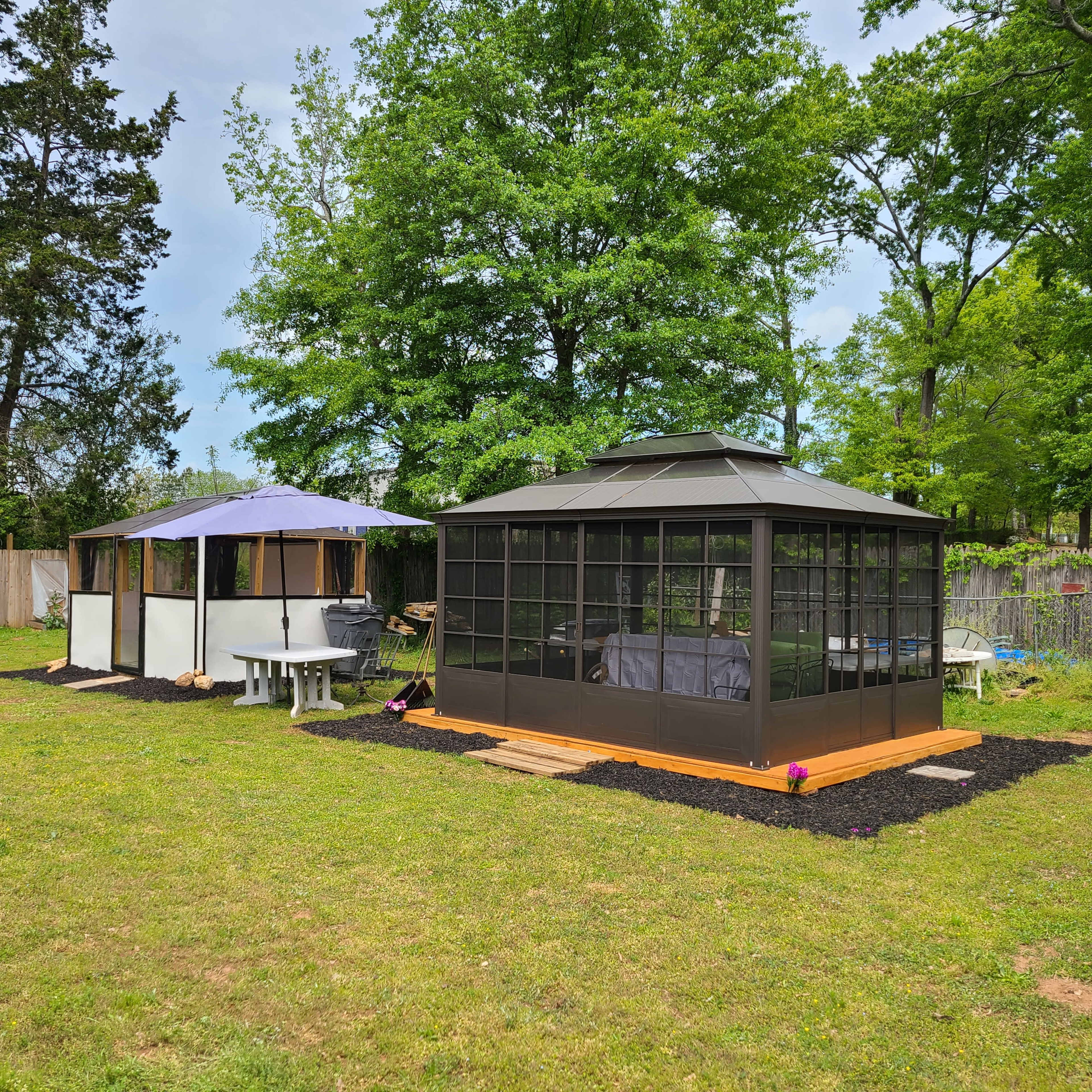 A black metal gazebo with glass walls sits beside a white canopy structure on a grassy area surrounded by trees.