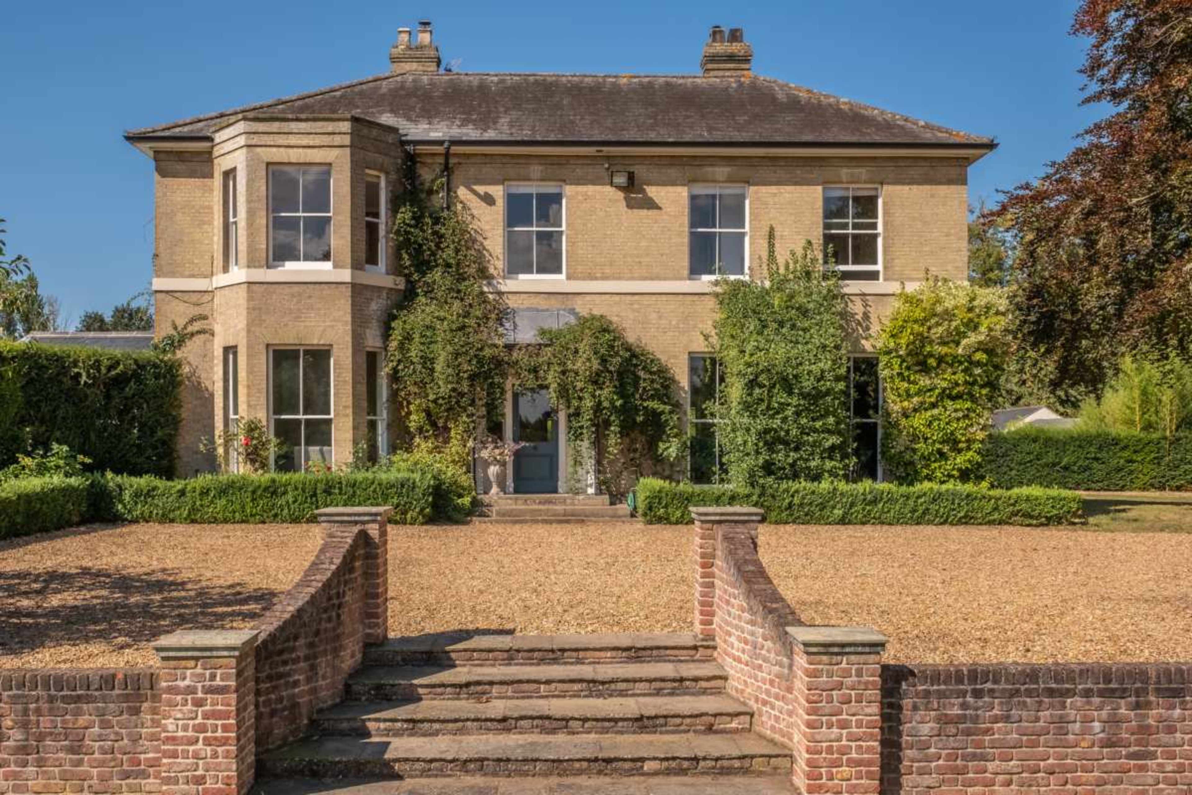 A two-story brick house with large windows is surrounded by manicured greenery and a gravel pathway leading up to the entrance.