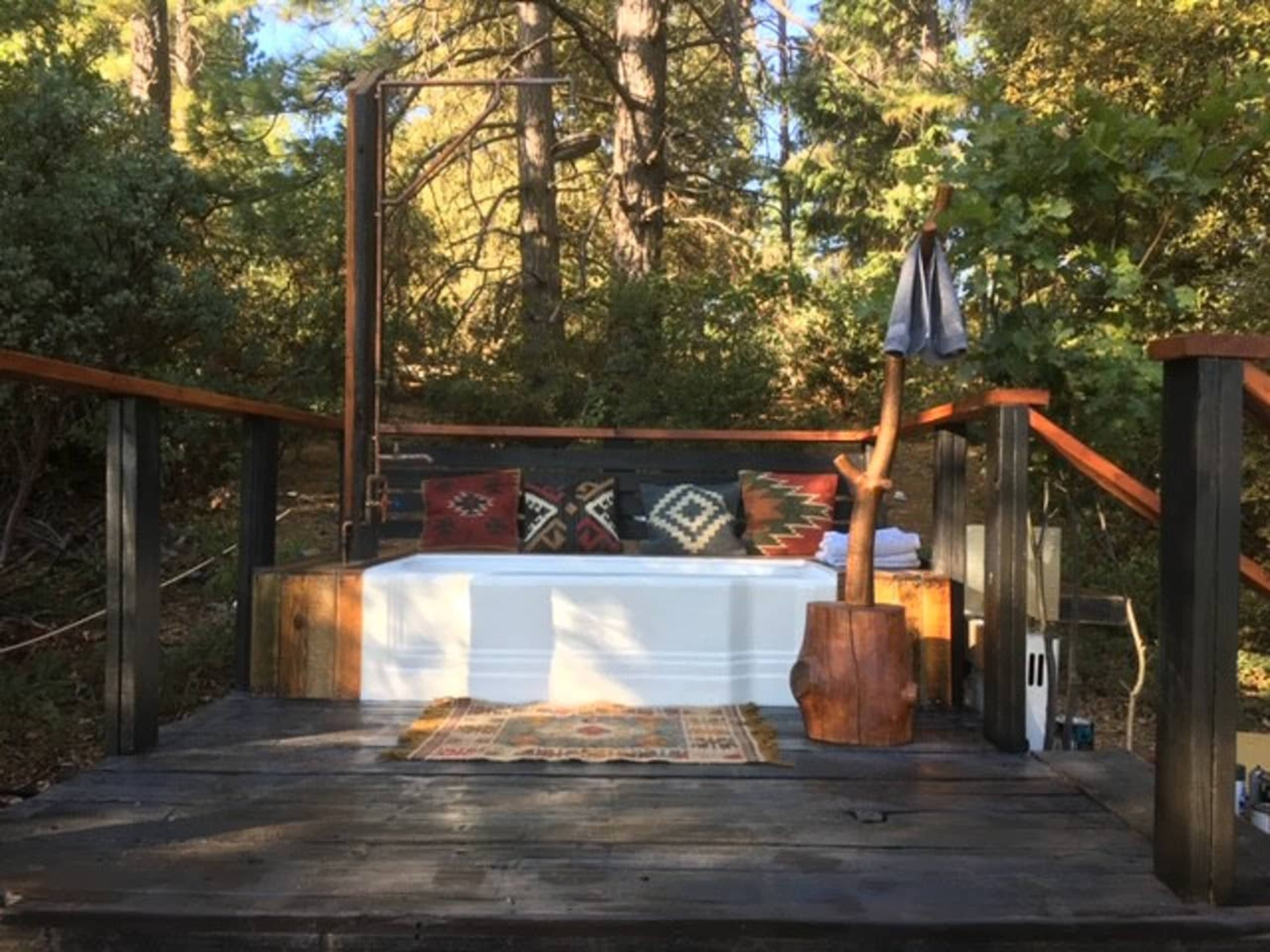 A wooden deck with a white tub surrounded by trees, decorative pillows, and a towel hanging from a pole.