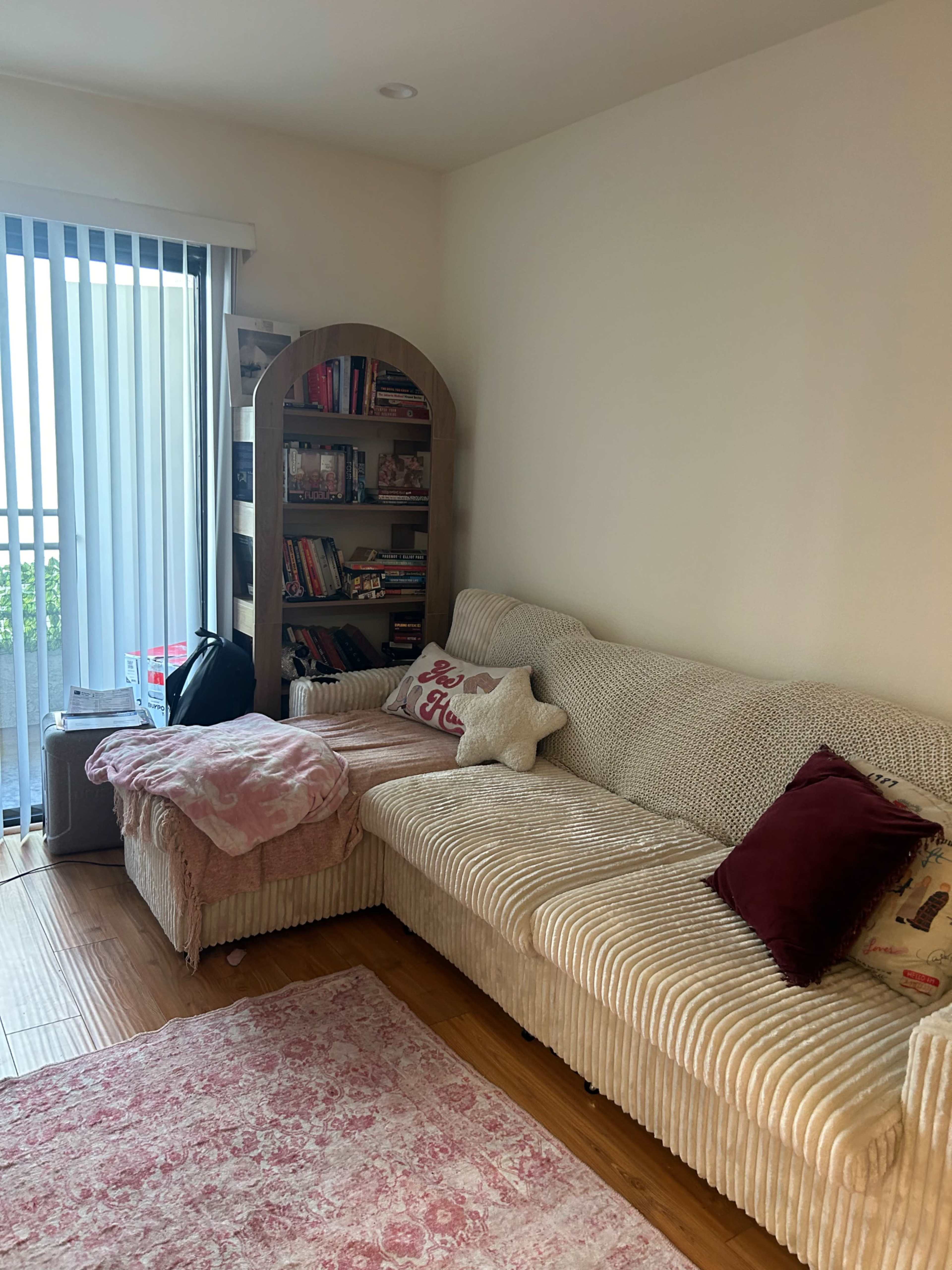The image shows a cozy living room featuring a light-colored sectional sofa, a small bookshelf filled with books, a pink throw blanket on the sofa, and a patterned rug on wooden flooring.