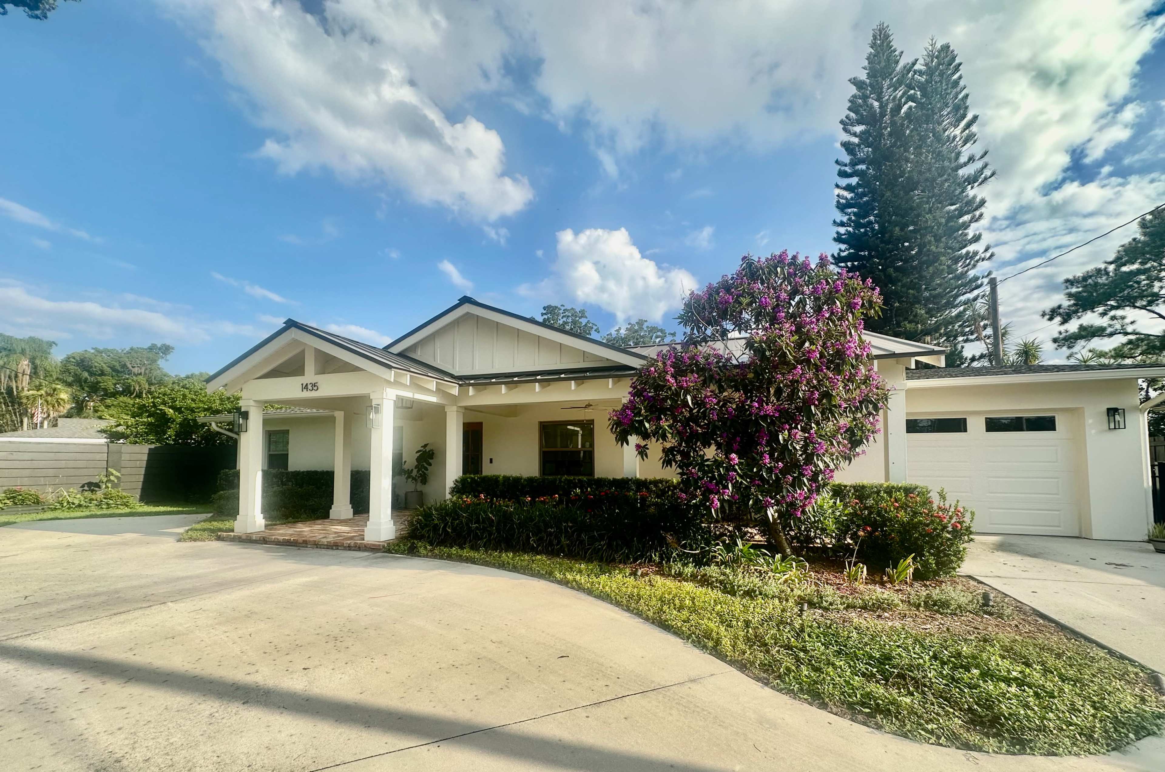 The house features a white exterior with a covered porch, surrounded by greenery and a flowering bush, set against a blue sky.