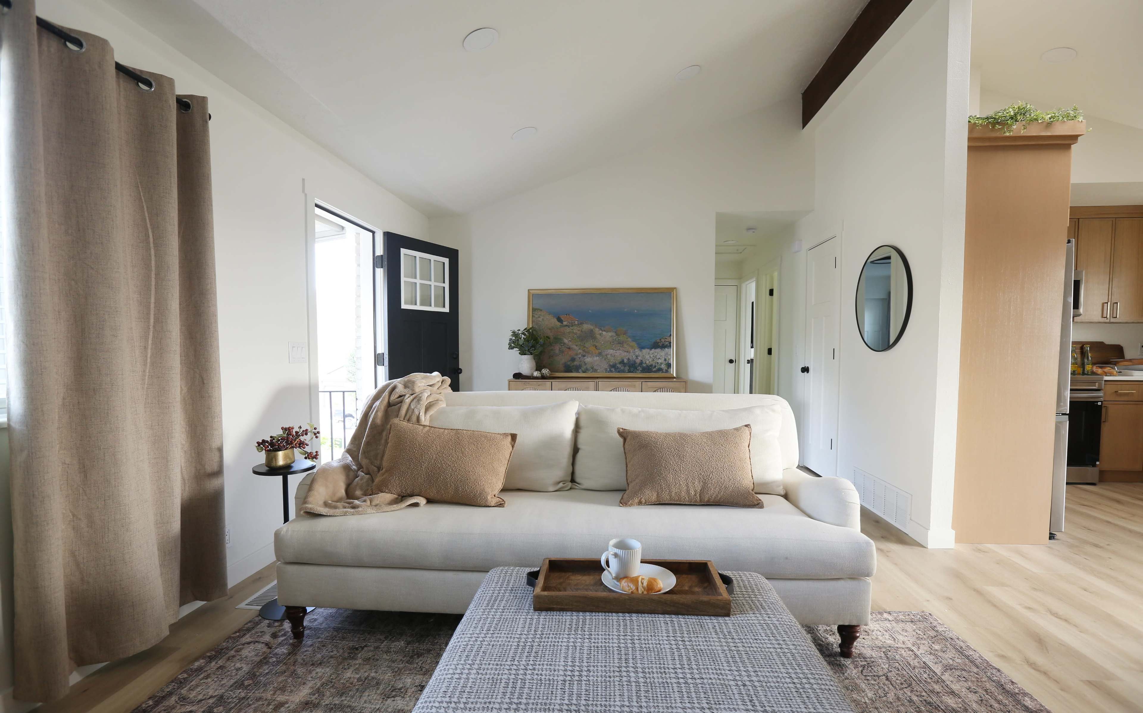 A cozy living room features a beige sofa with decorative pillows, a wooden coffee table, and a painting on the wall, with natural light coming through a door.