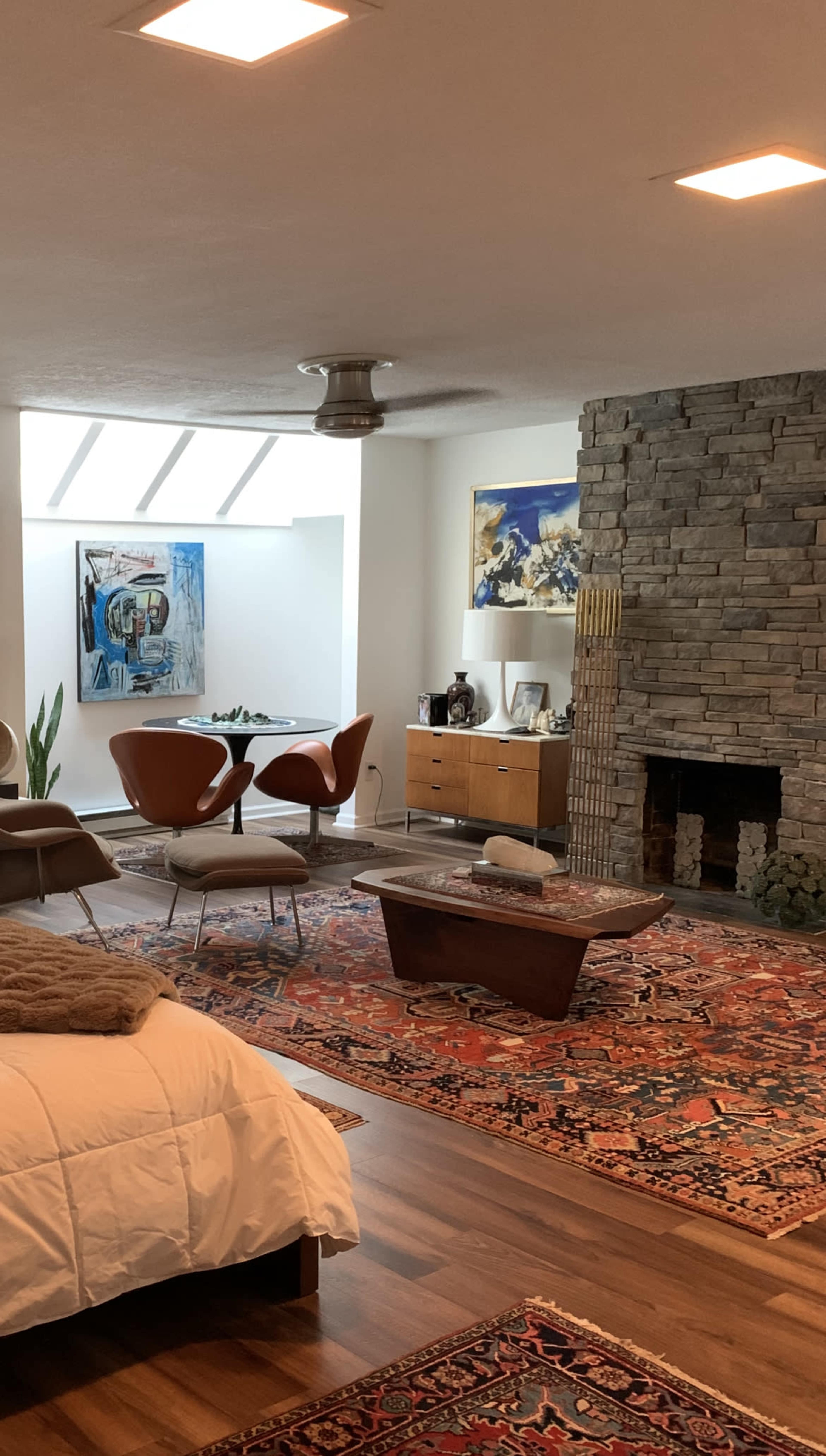 The image shows a modern living space with a bed in the foreground, a stone fireplace, a wooden coffee table, and a dining area featuring a round table and chairs.