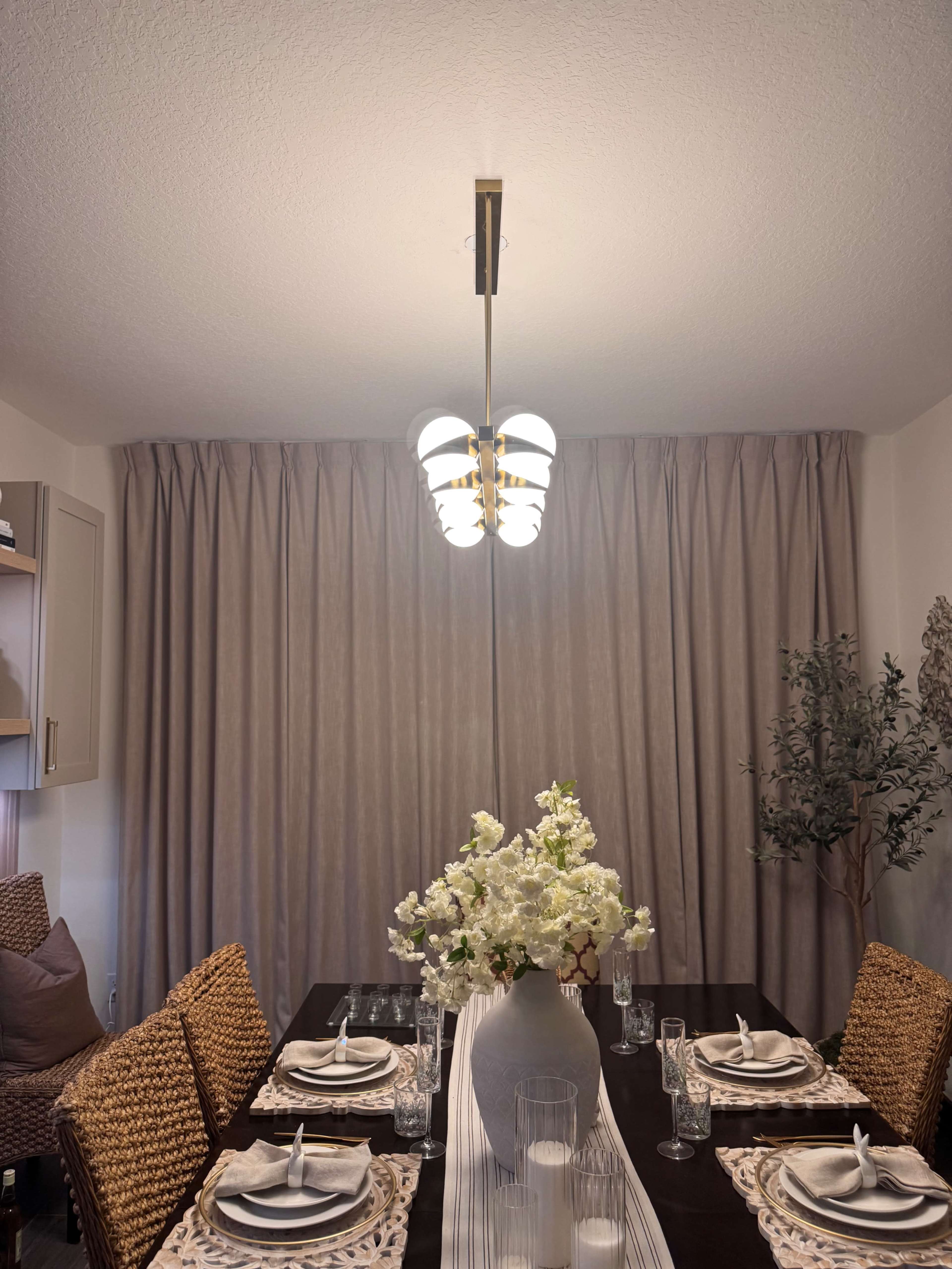 A dining table is set with plates and glasses, topped with a white vase of flowers, under a modern light fixture and surrounded by woven chairs.