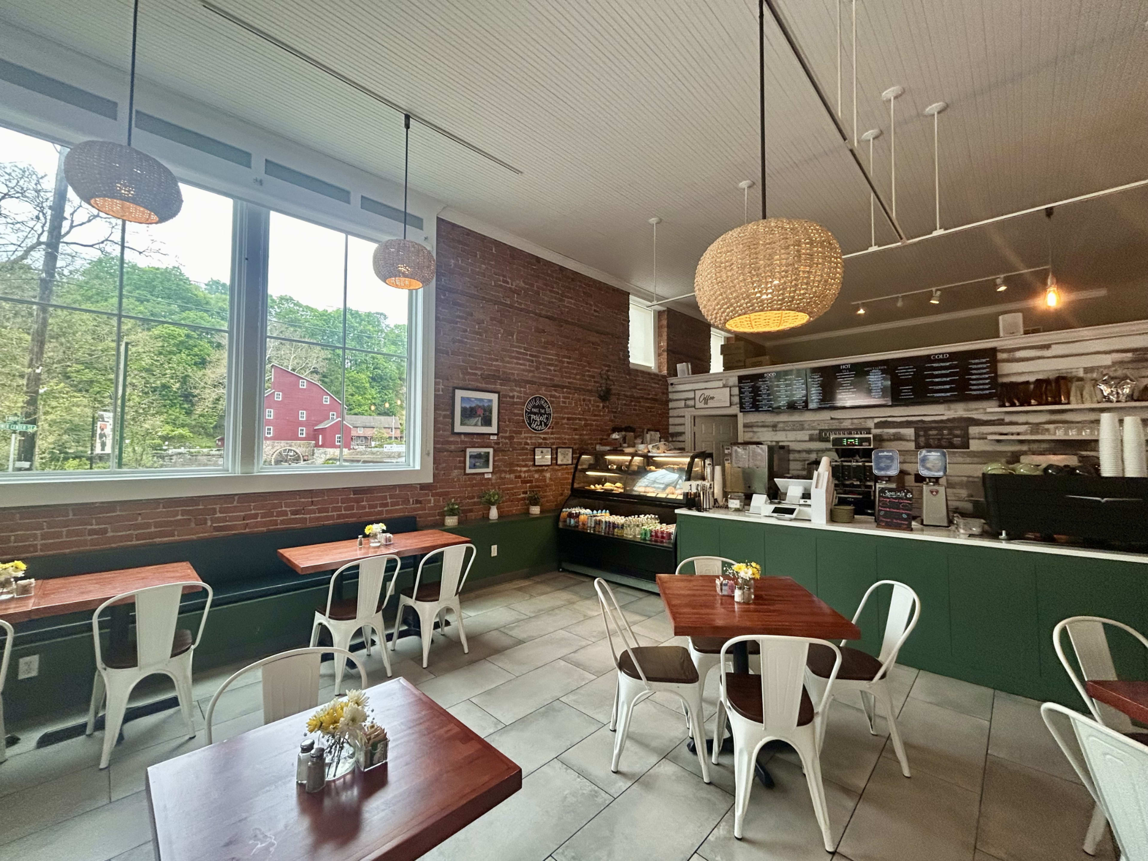 The image shows a spacious café with wooden tables and chairs, large windows allowing natural light in, and a counter displaying various food items.