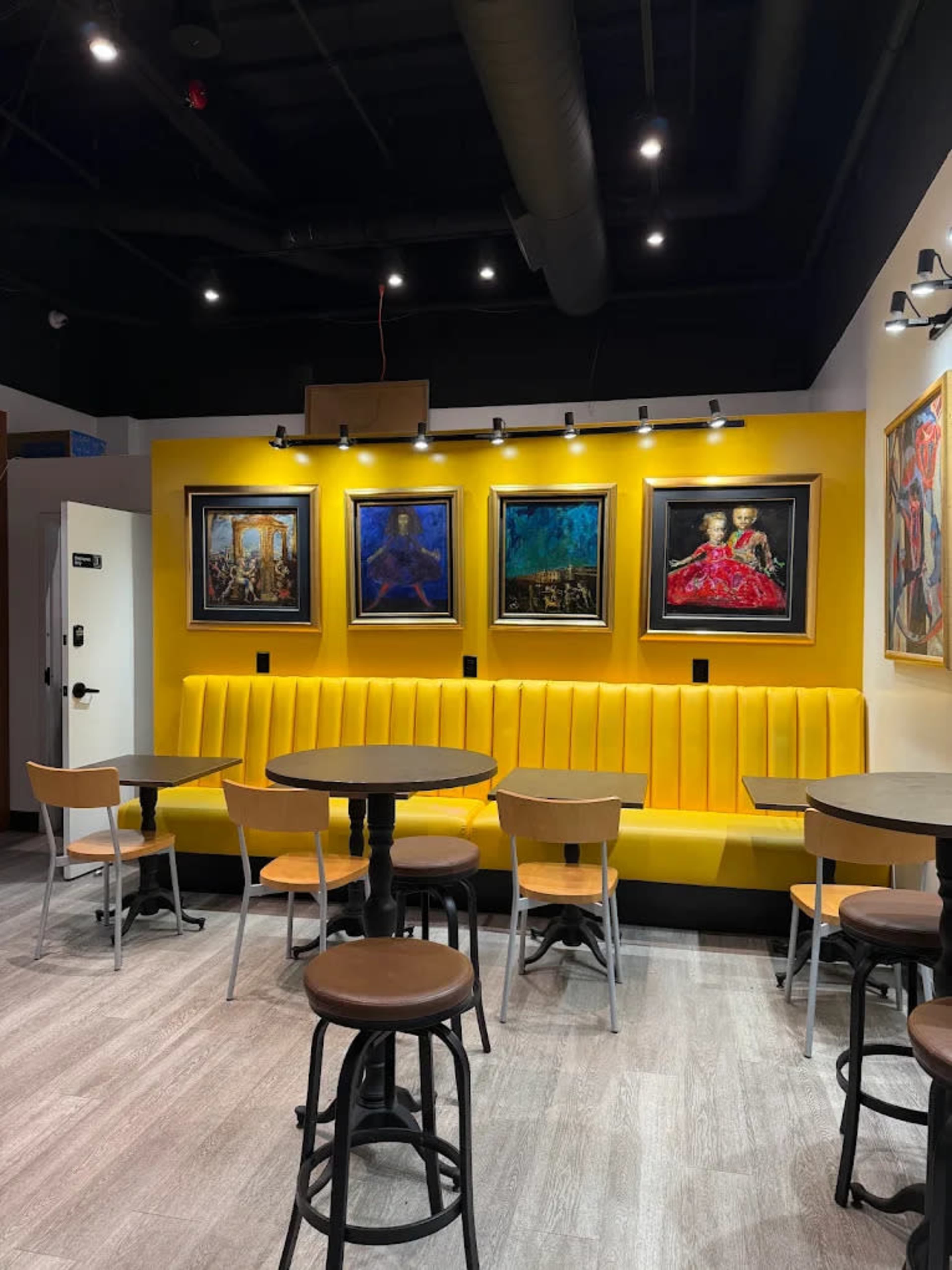 The image shows a cafe interior with yellow upholstered seating, round tables, and framed paintings on the wall.