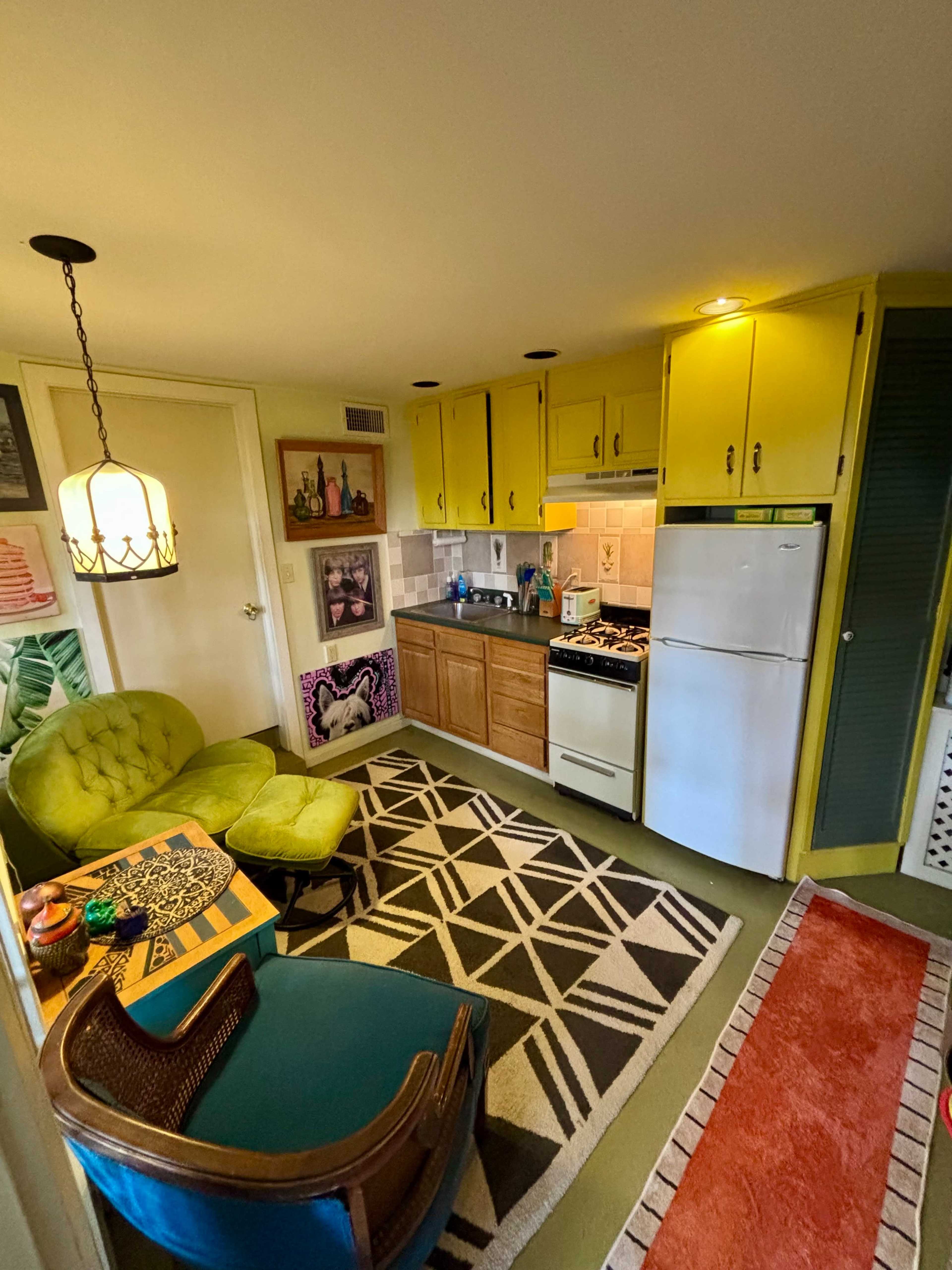 The image shows a small kitchen area featuring yellow cabinets, a white refrigerator, and a cozy seating arrangement with colorful chairs and a geometric rug.