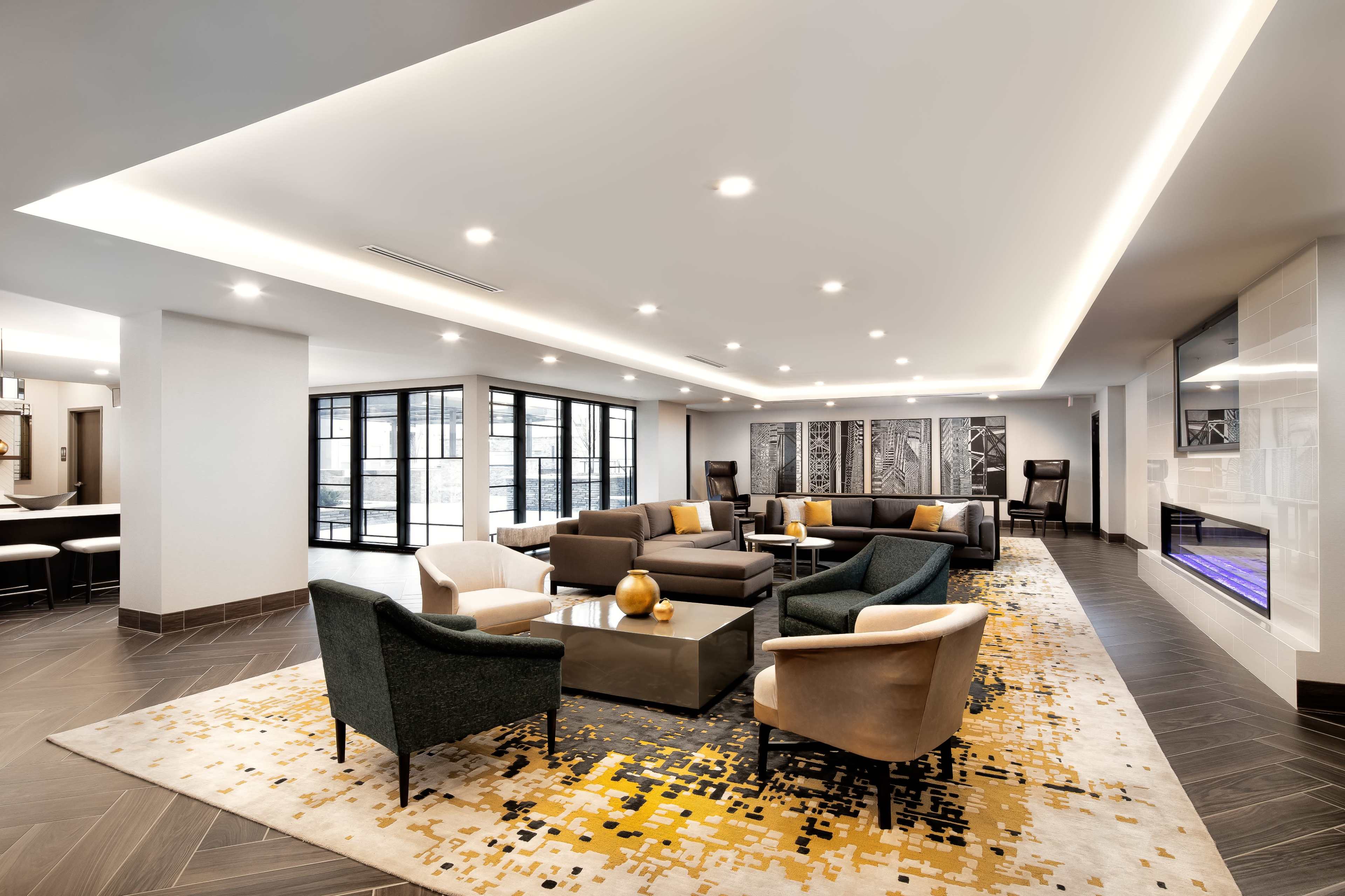 The image shows a modern lounge area with comfortable seating, a large area rug, and ambient lighting.