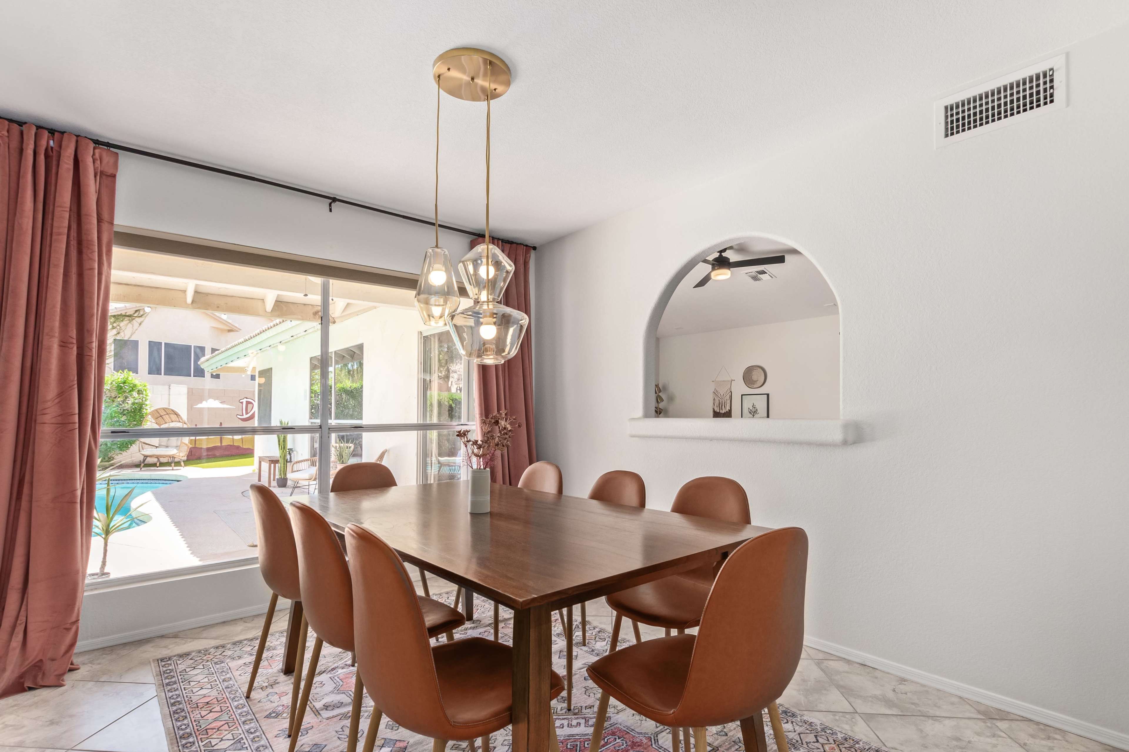 A dining area features a wooden table surrounded by eight brown chairs, with a large window showcasing an outdoor space and a pendant light hanging above.