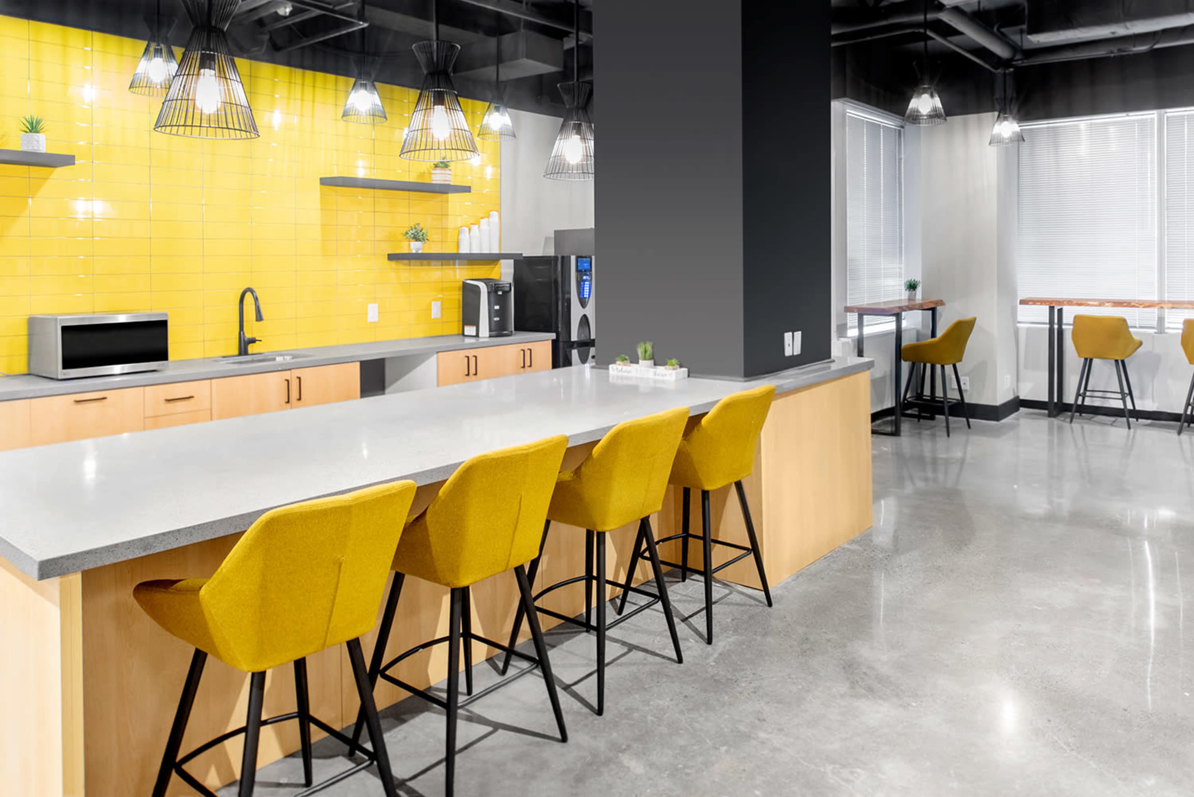 The image shows a modern kitchen space featuring a large gray countertop with yellow bar stools, a yellow tiled wall, and various kitchen appliances.