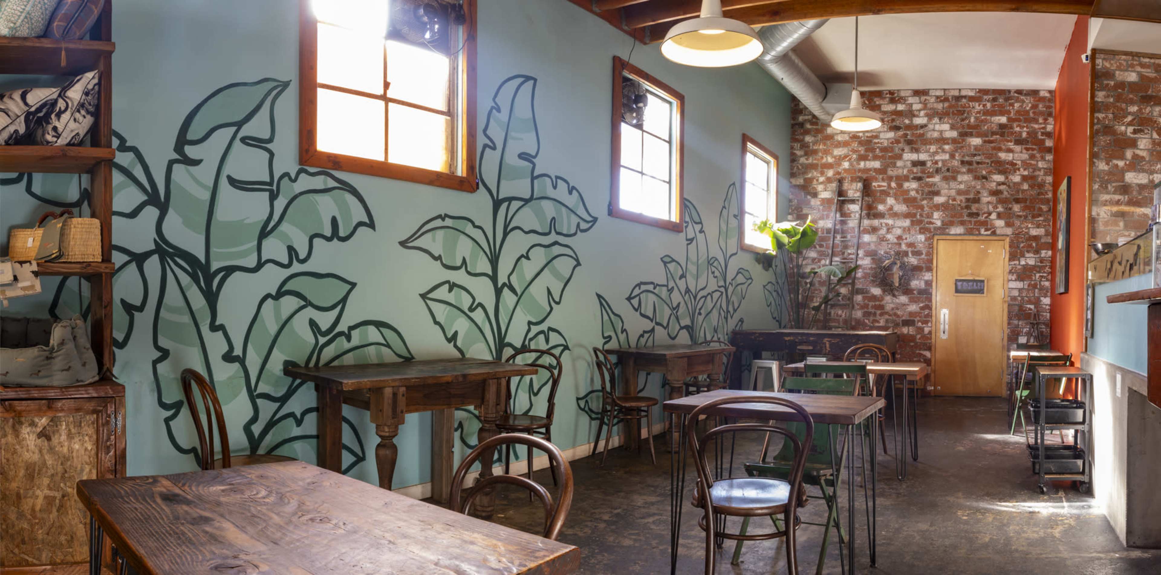 The interior of a cafe features wooden tables, a brick wall, and murals of large green leaves on the blue walls.