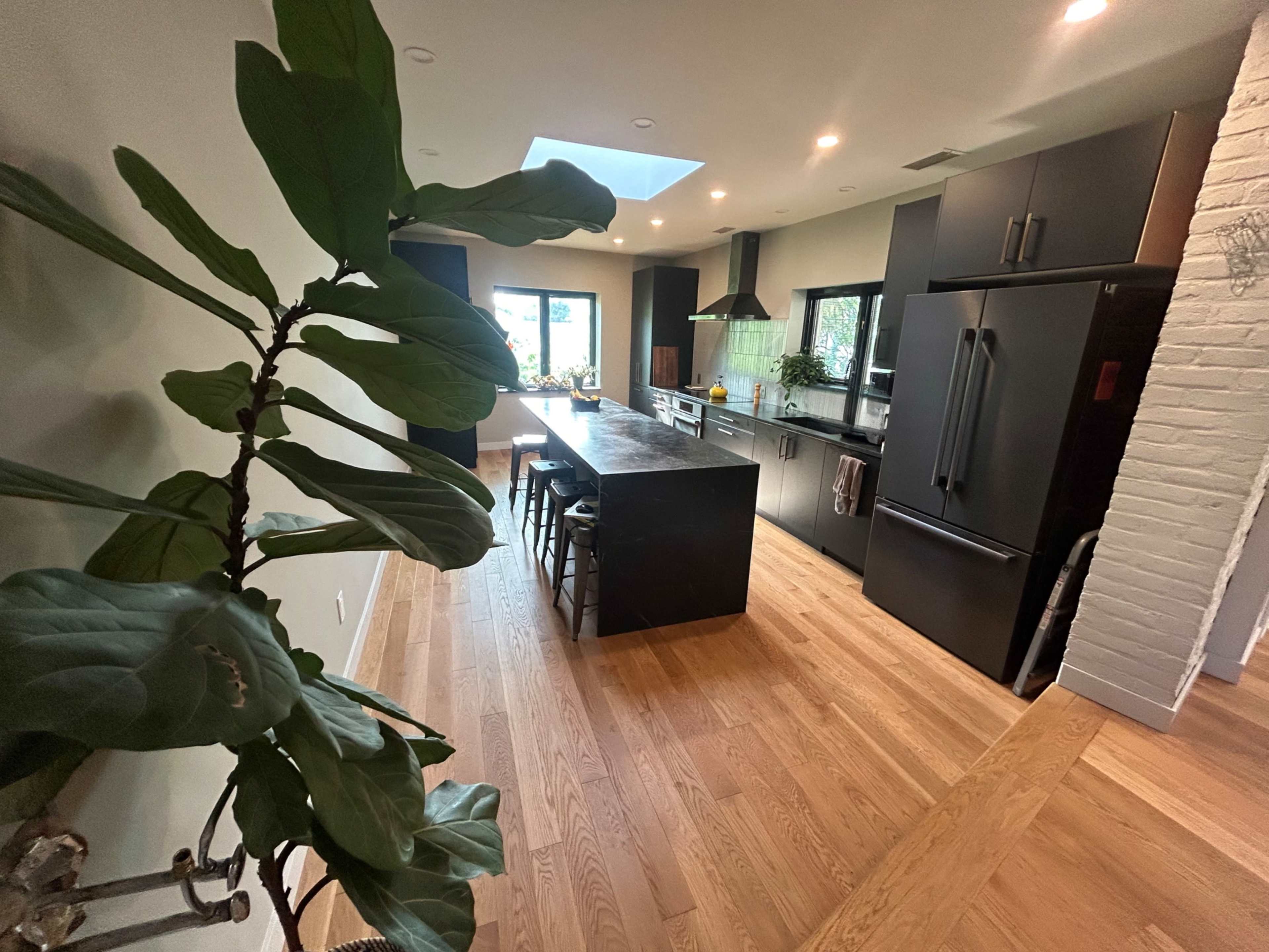 The image shows a modern kitchen with dark cabinetry, a central island with seating, and large windows allowing natural light to enter.