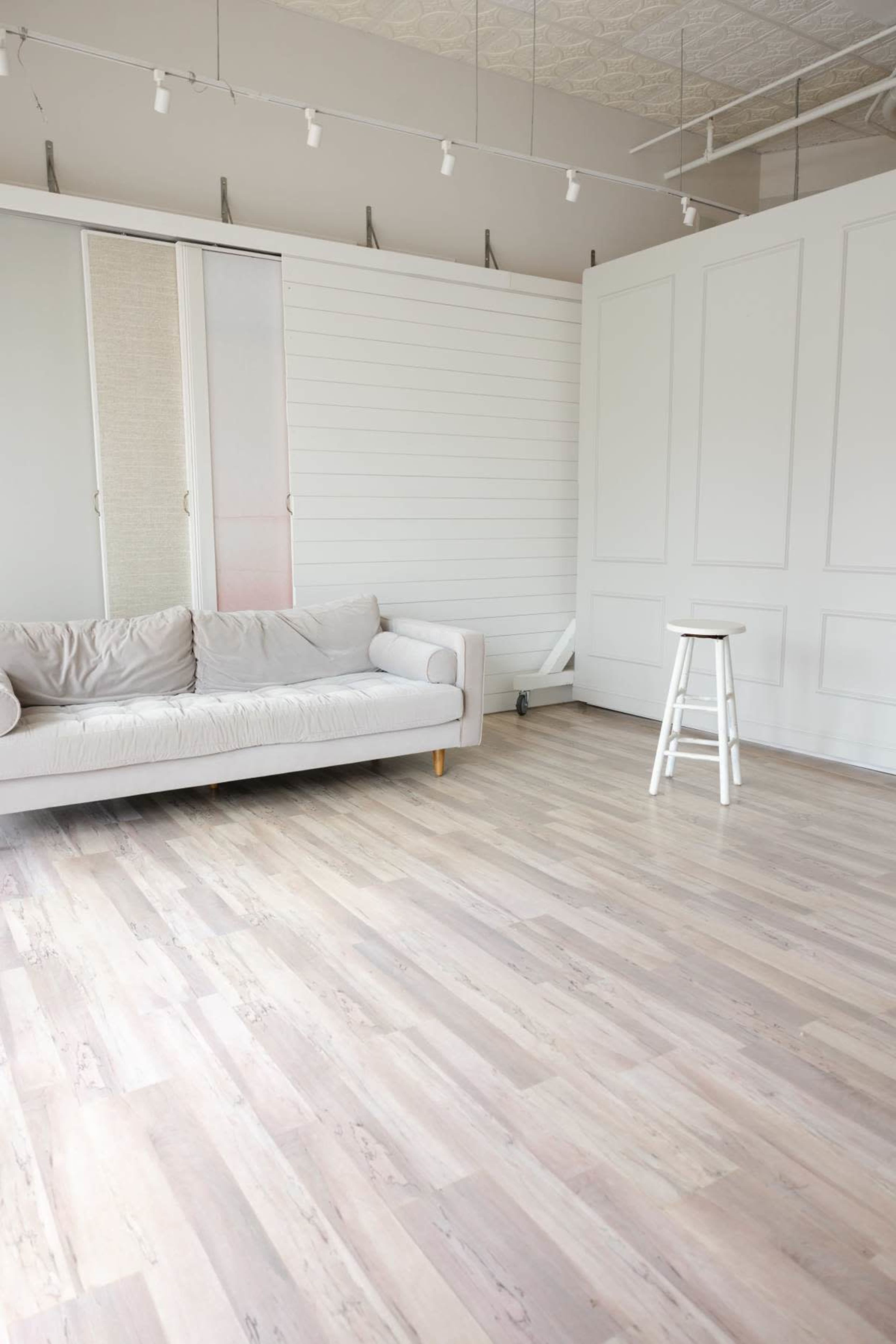 A light-filled room features a beige sofa against a white wall with a stool and wood laminate flooring.