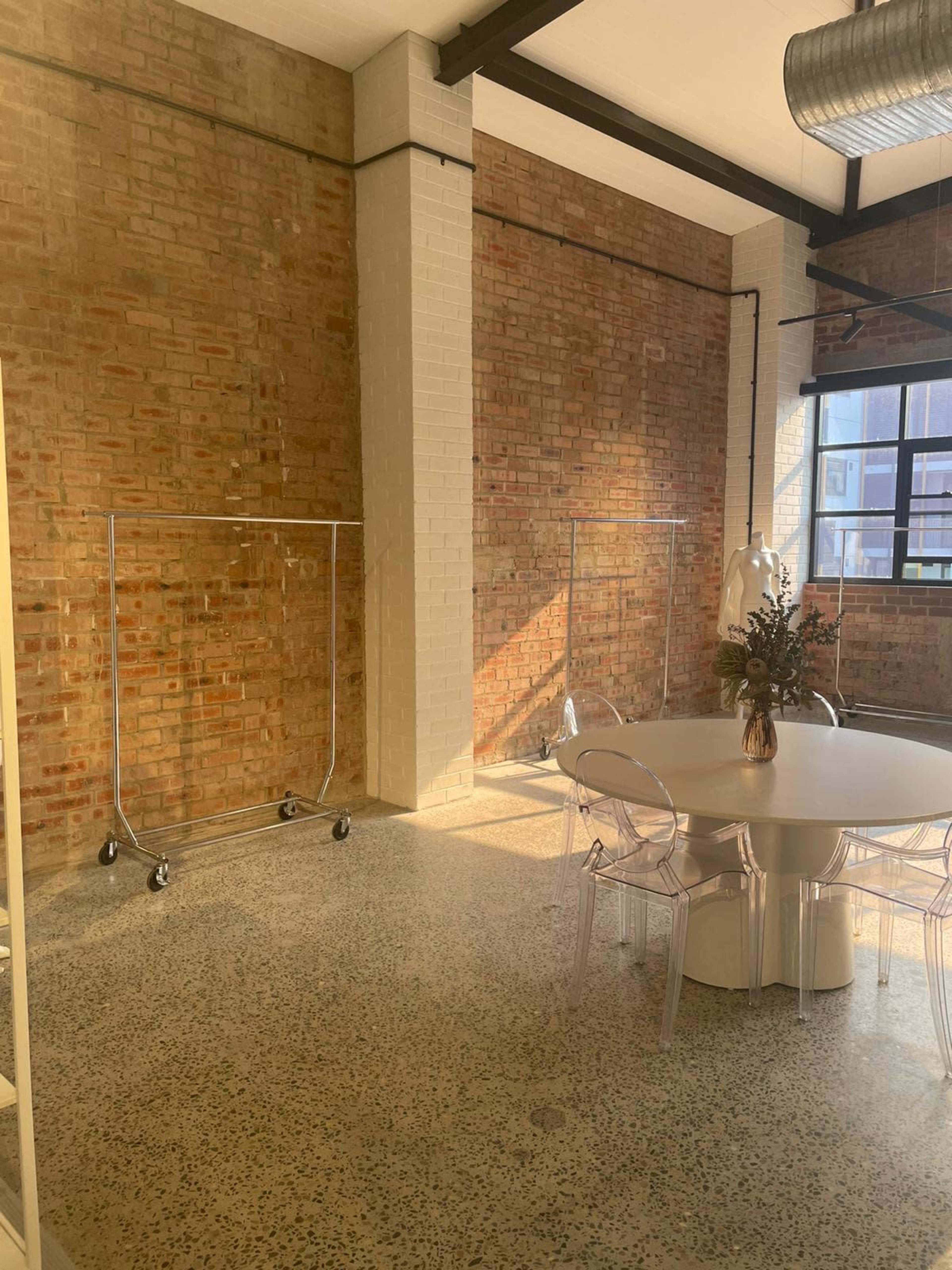 A bright, modern interior space featuring two clothing racks, a round table with floral decor, and exposed brick walls.