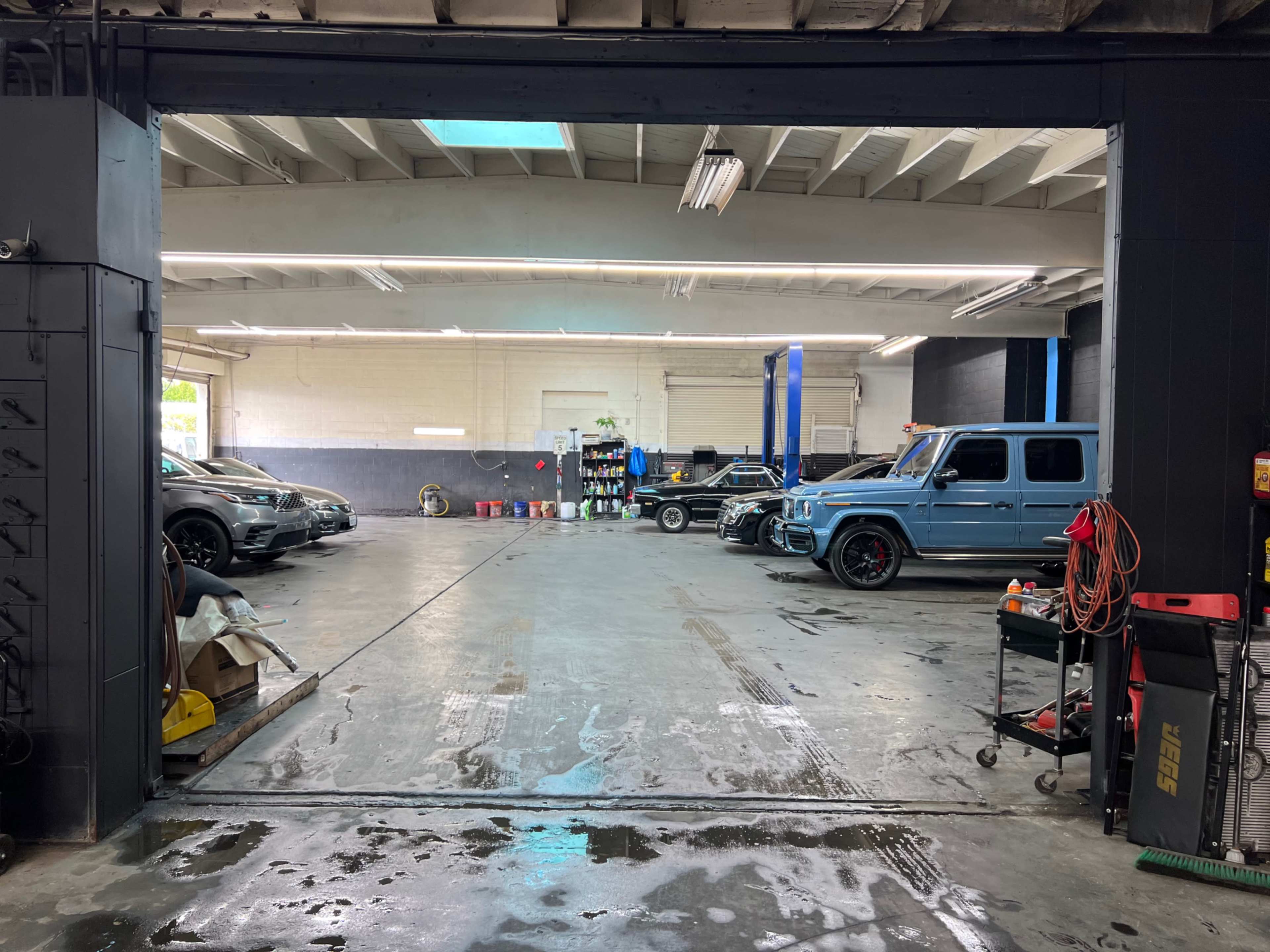 The image shows an automotive workshop with several vehicles parked inside, tools and supplies organized along the walls, and a lift in the background.