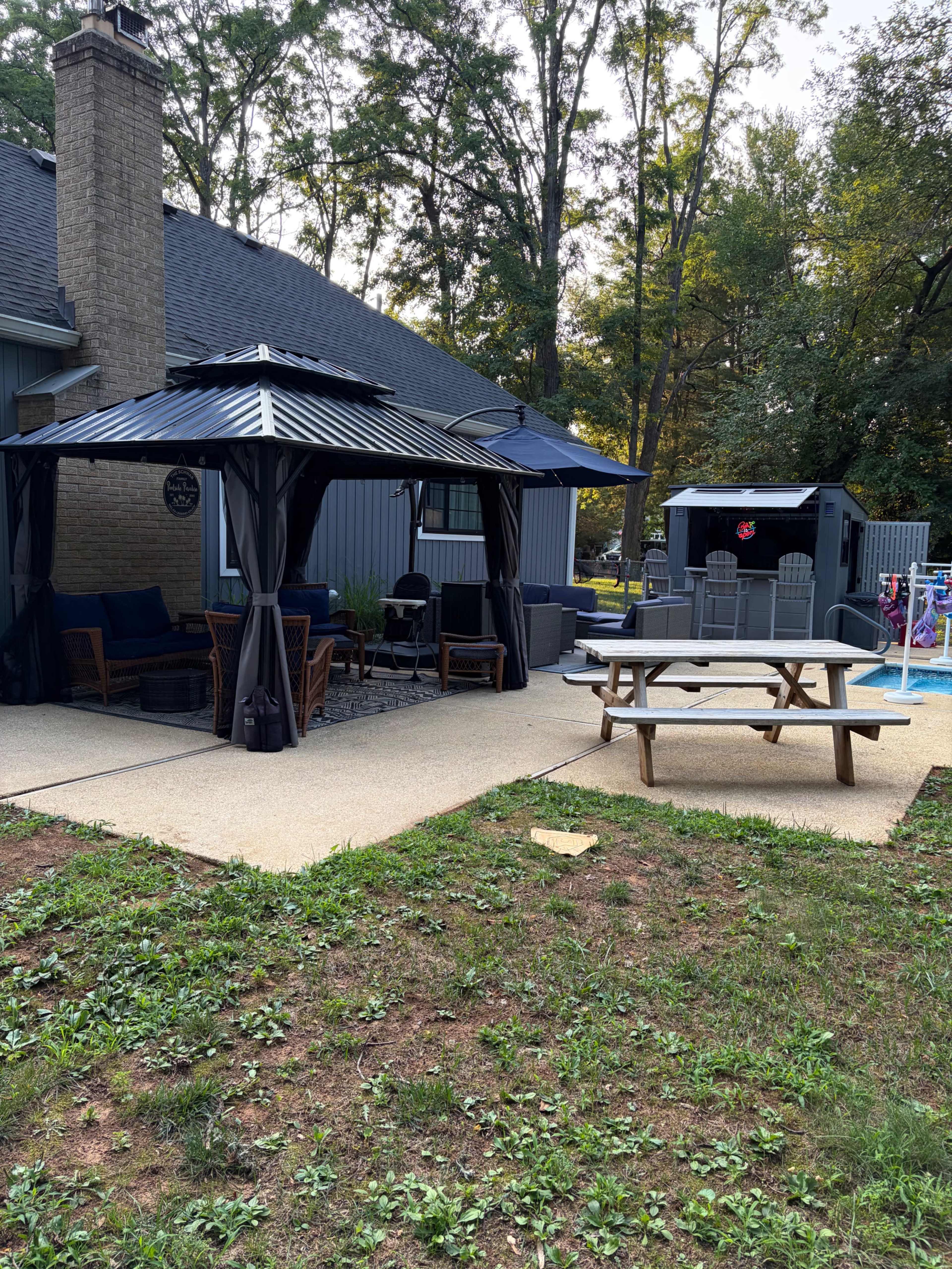 The image shows a backyard with a gazebo, a picnic table, and a pool area surrounded by trees.