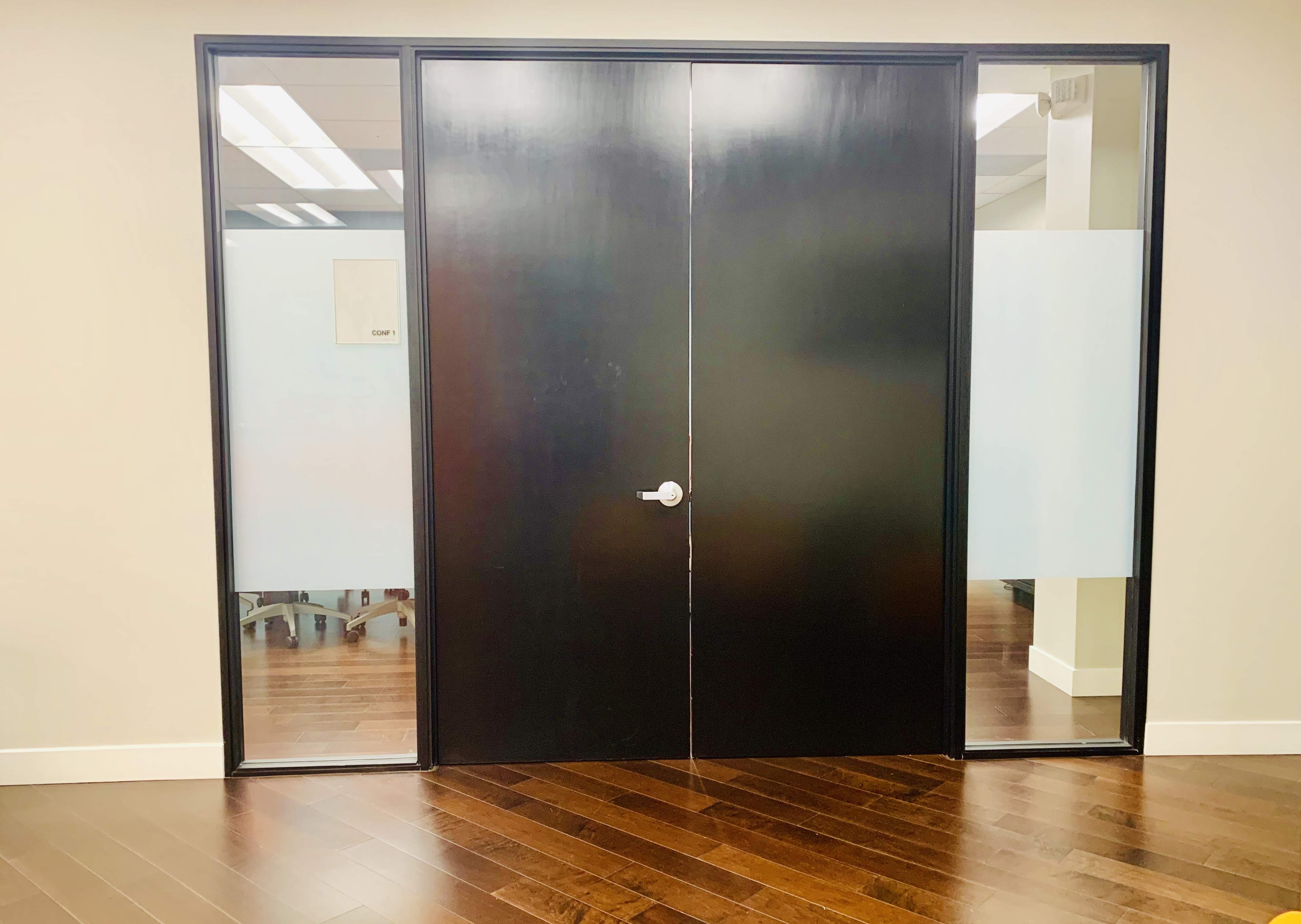 A pair of large black double doors framed by glass panels, set against a wall with wooden flooring.