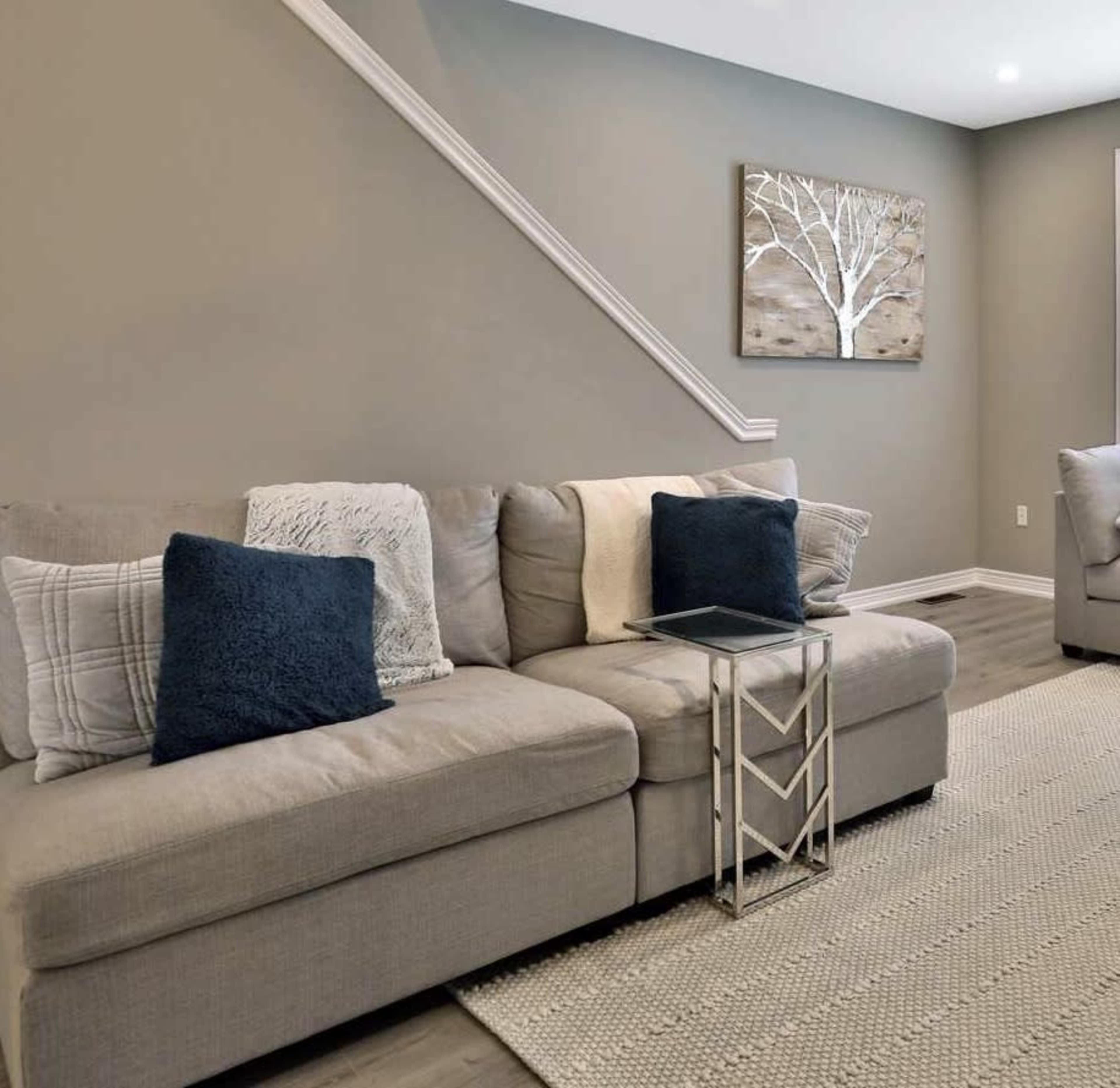 A gray sectional sofa with pillows sits beside a small glass side table on a textured rug in a room with light gray walls and artwork featuring a tree.