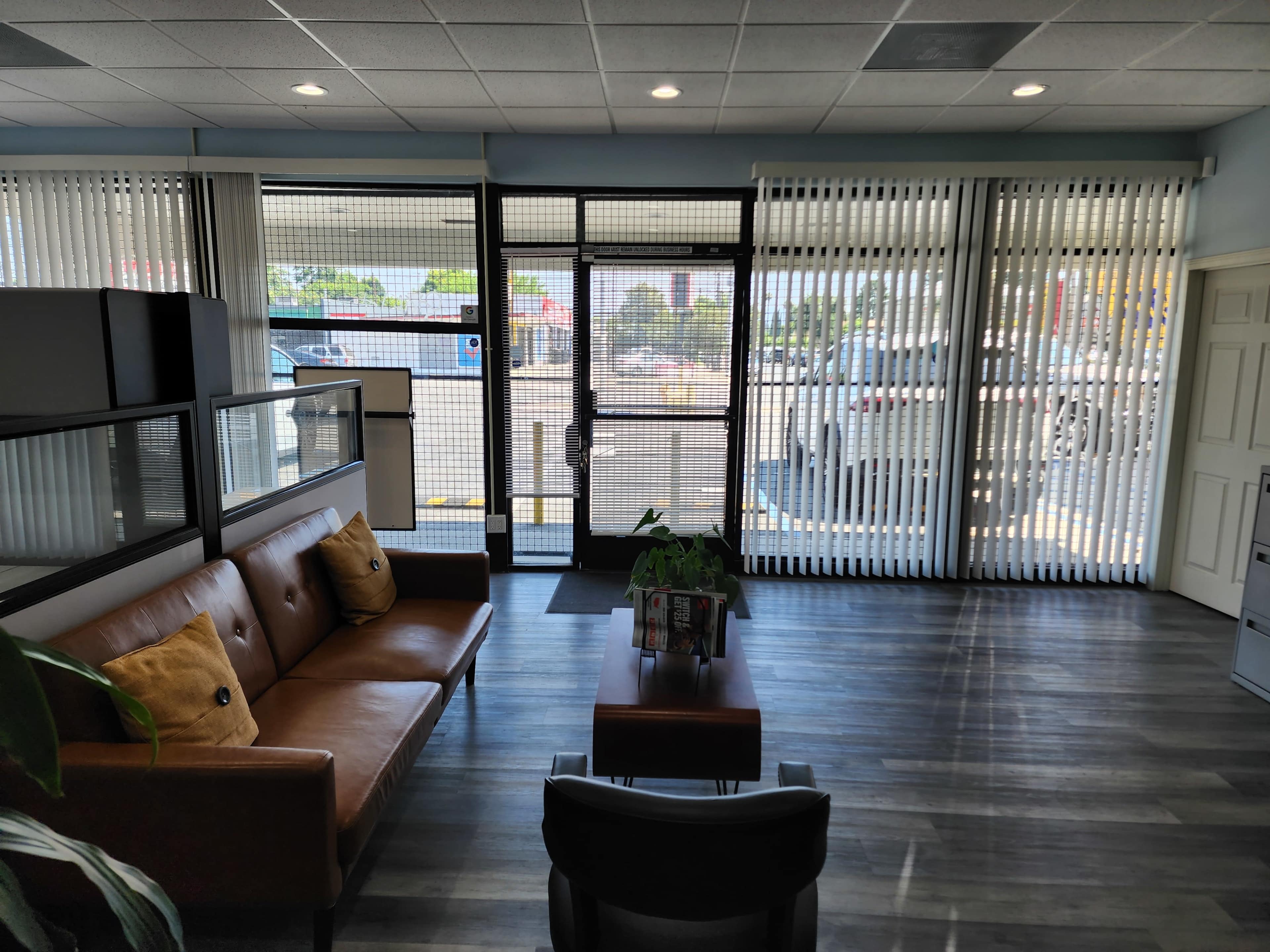 The image shows a modern waiting area with a brown leather sofa, a coffee table, and large windows covered with vertical blinds.