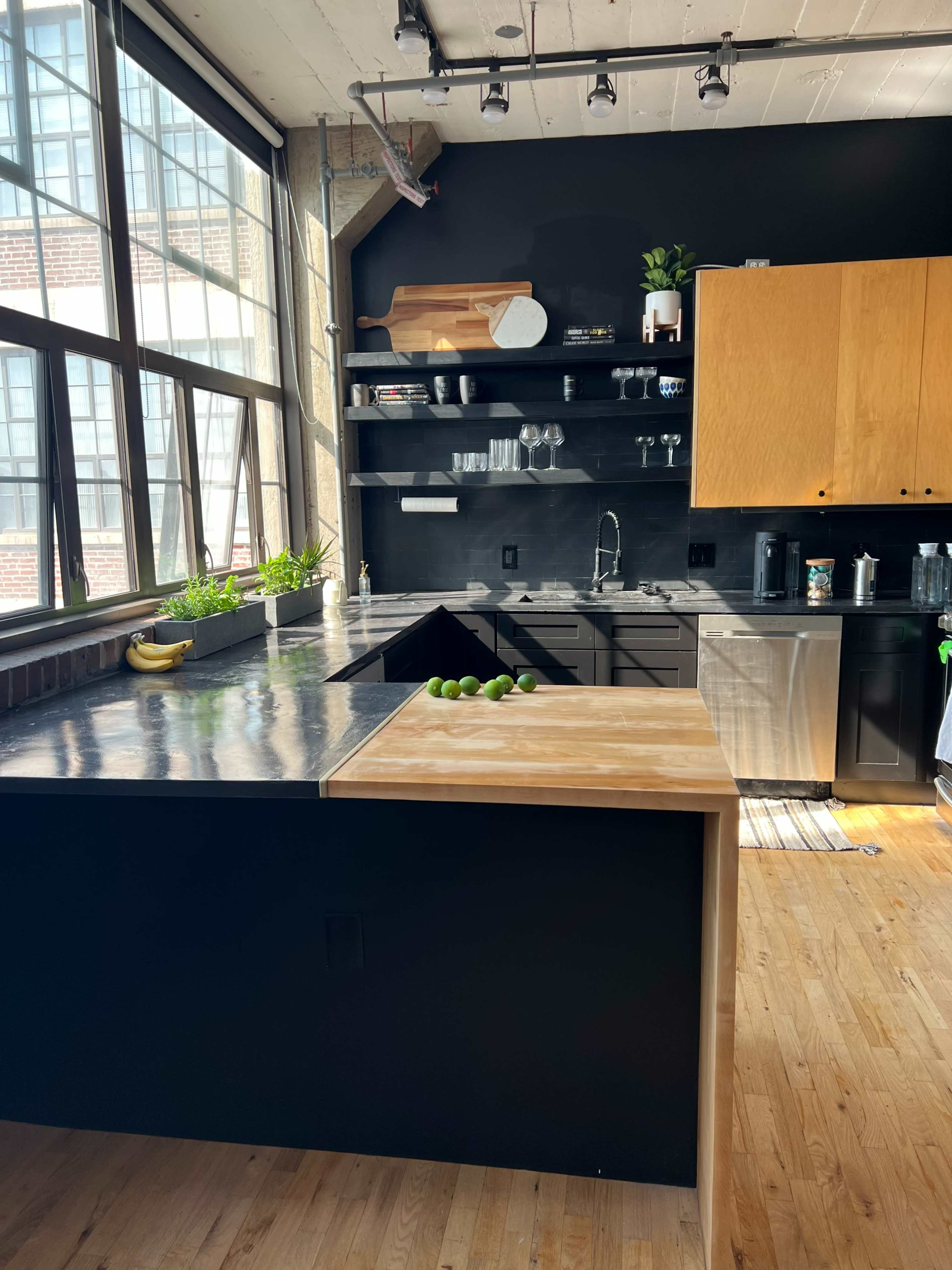 A modern kitchen with a black and wood color scheme, featuring large windows, open shelving, and stainless steel appliances.