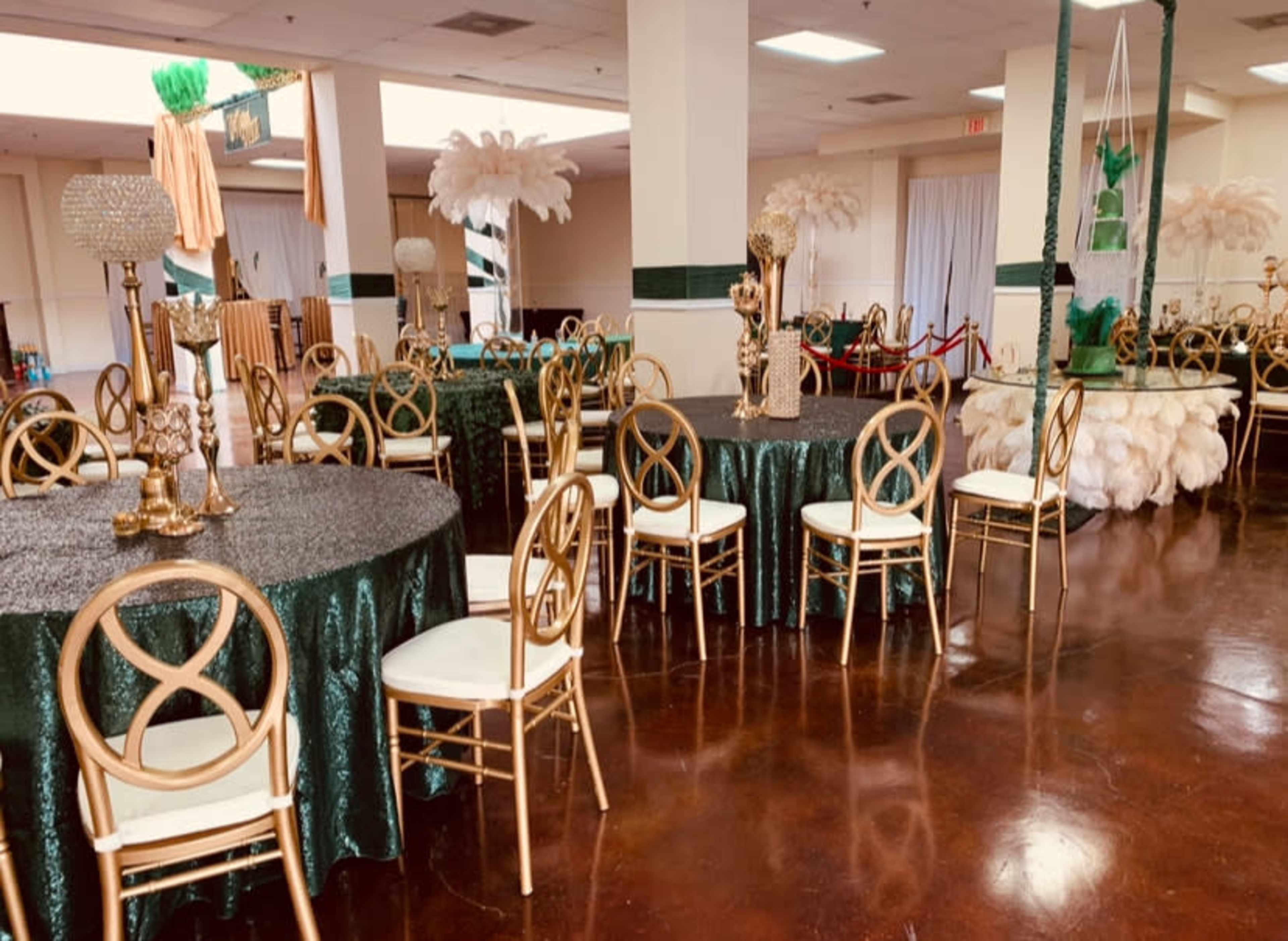 The image shows a decorated banquet hall with round tables covered in dark green tablecloths and gold chairs, featuring elegant centerpieces and large feather arrangements in the background.