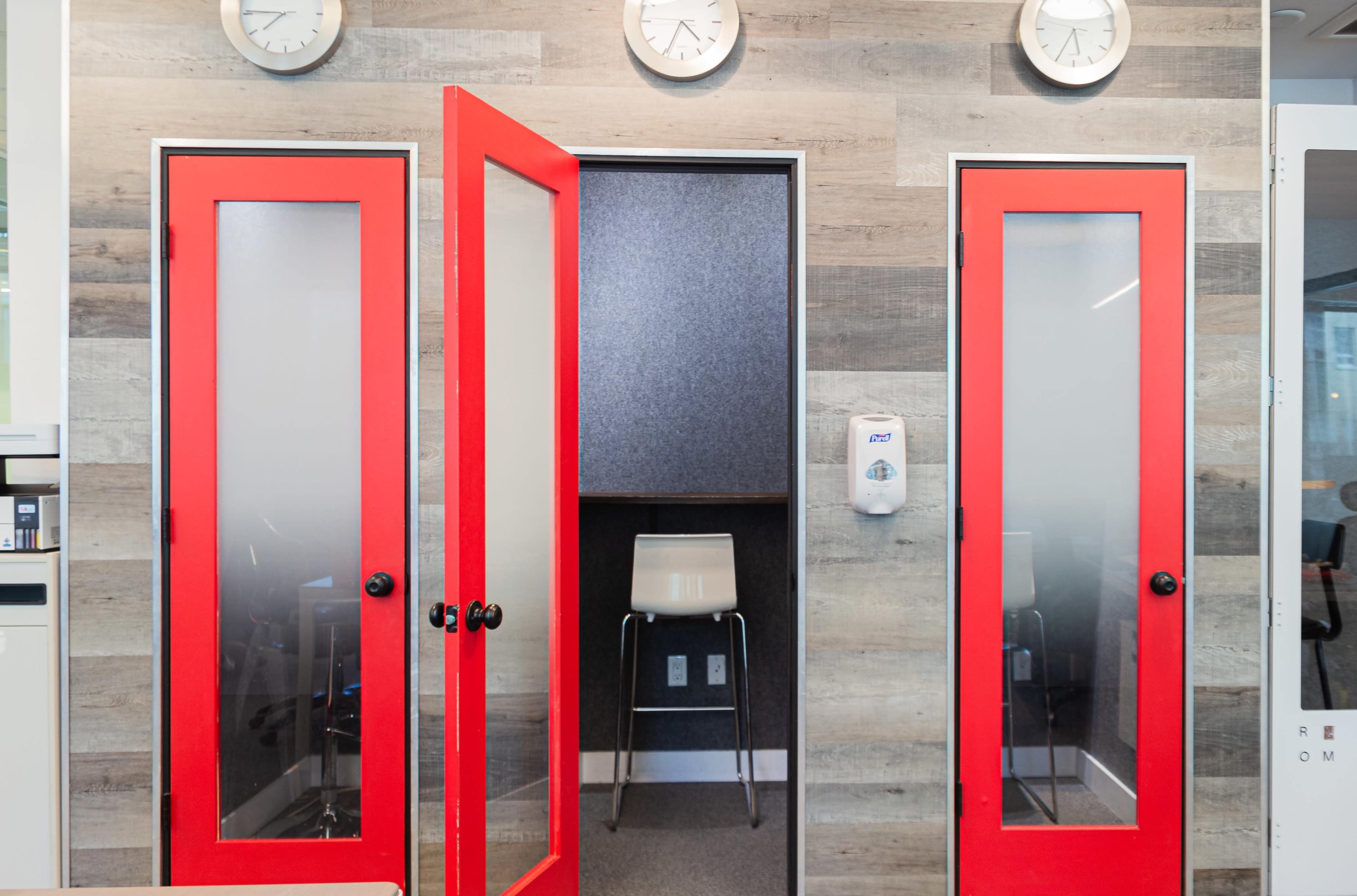 A modern office space featuring three red doors with frosted glass, leading to a private area with a chair and a hand sanitizer dispenser.
