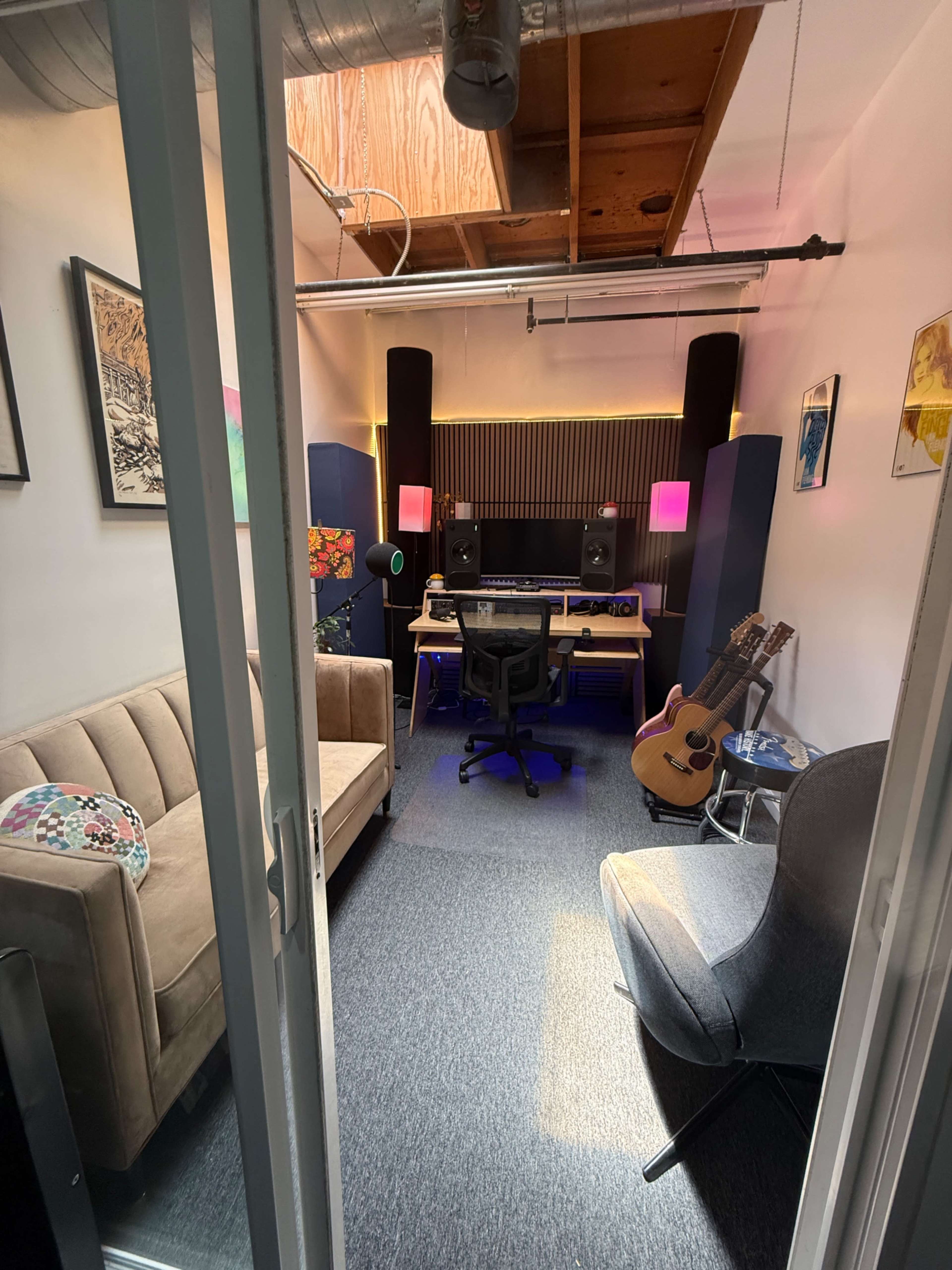 The image shows a modern music studio with a command desk, acoustic panels, a sofa, and a guitar positioned beside a gray chair.