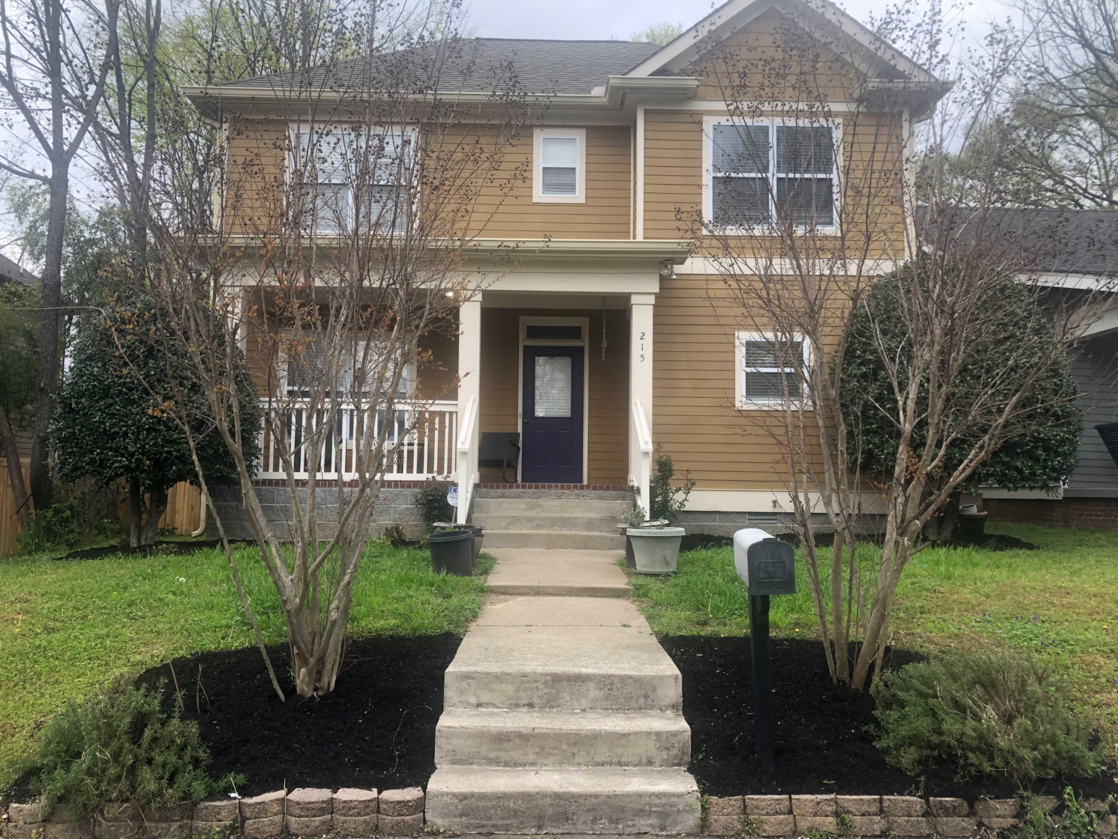 The image shows a two-story house with a front porch, flanked by bare trees and landscaped grass.