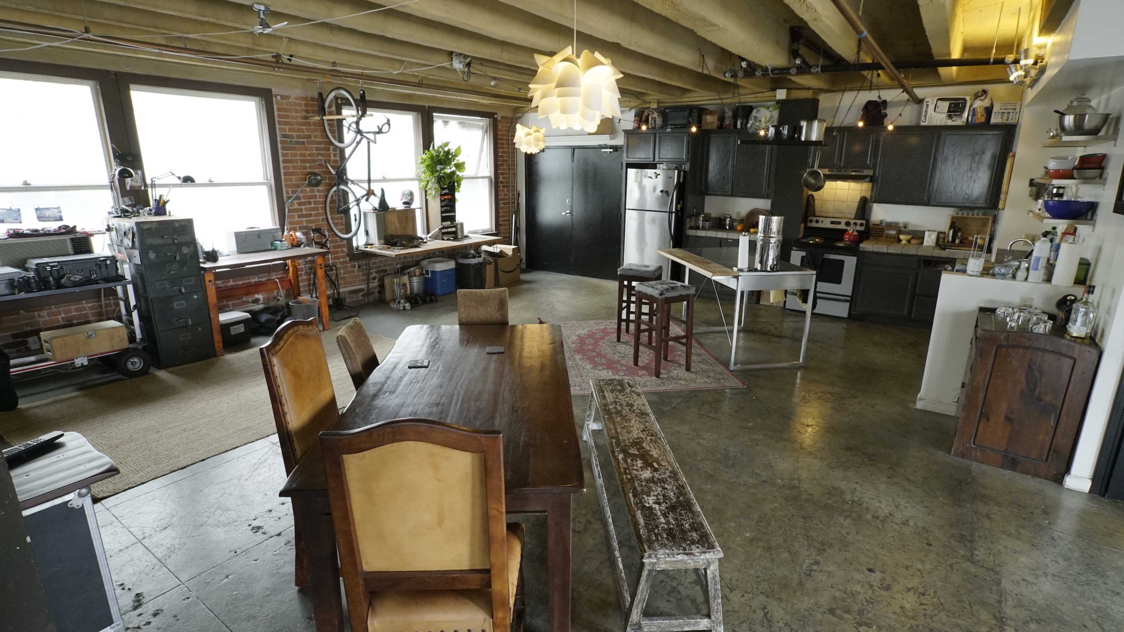 The image shows an open-concept kitchen and dining area featuring a wooden table, a bench, and modern appliances in a spacious, industrial-style loft.