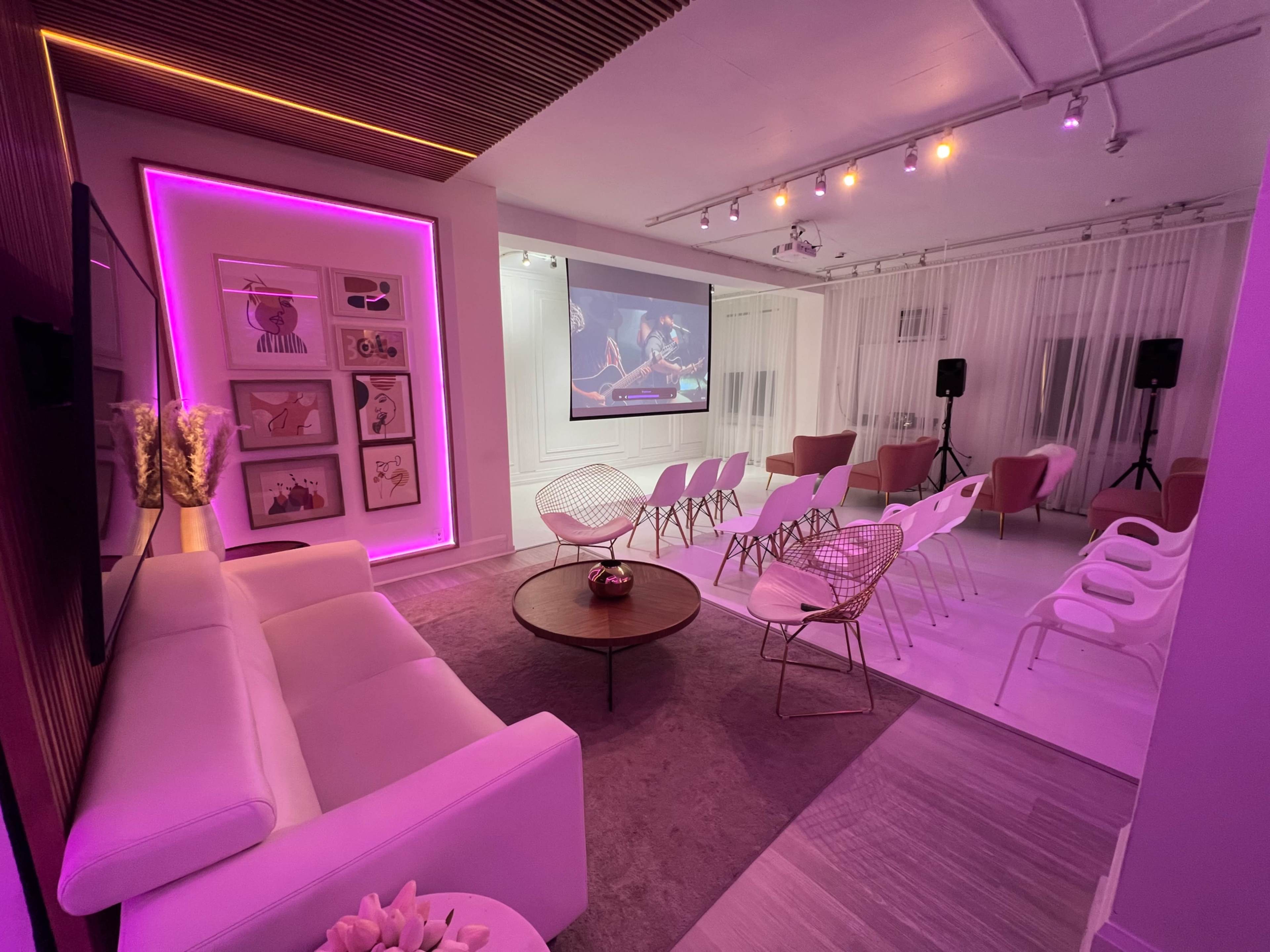 A modern event space featuring a white couch, a round coffee table, a large screen displaying an image, and rows of chairs under soft lighting.