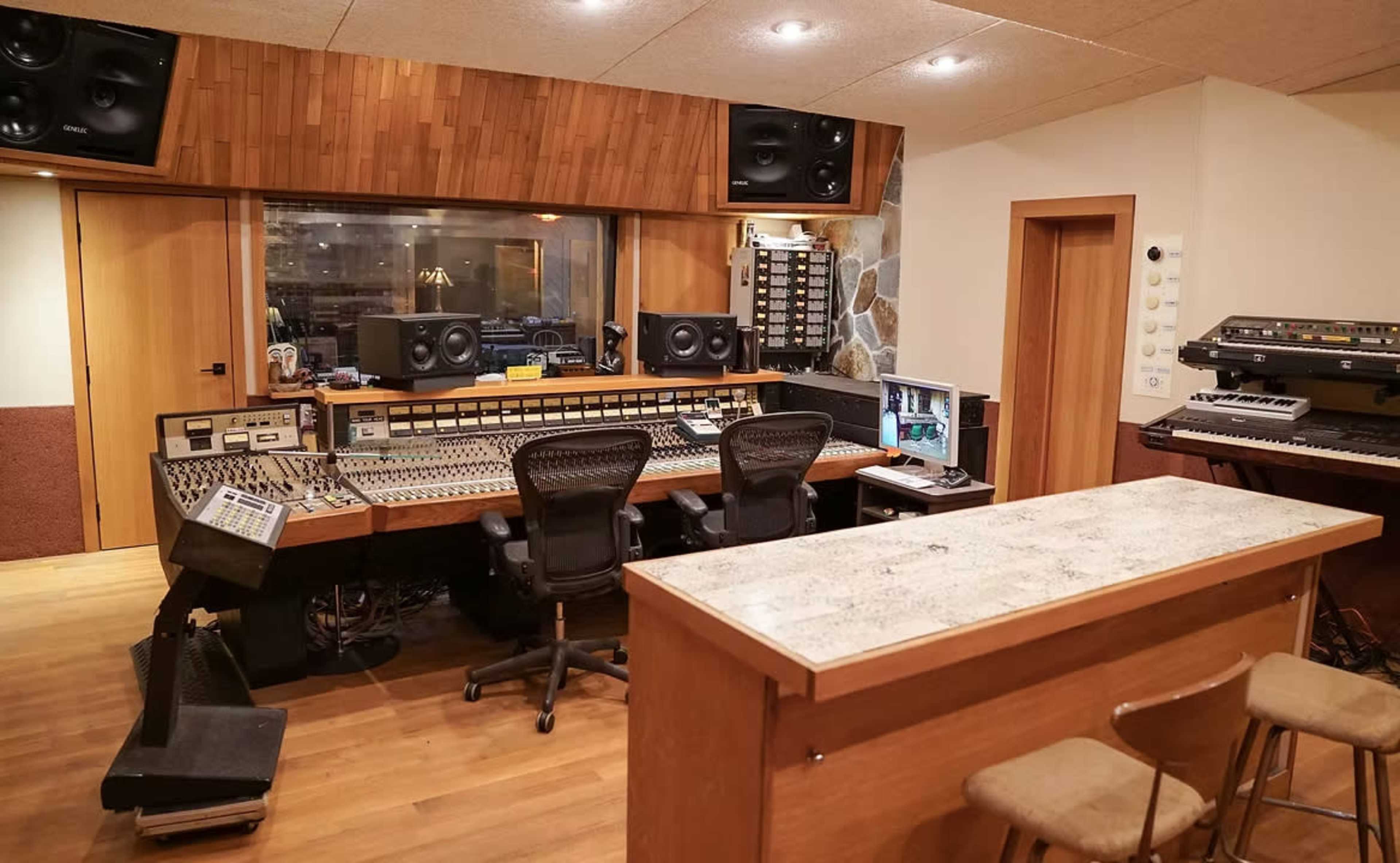 The image shows a recording studio with a large mixing console, multiple speakers, and various musical equipment set against wooden walls and flooring.
