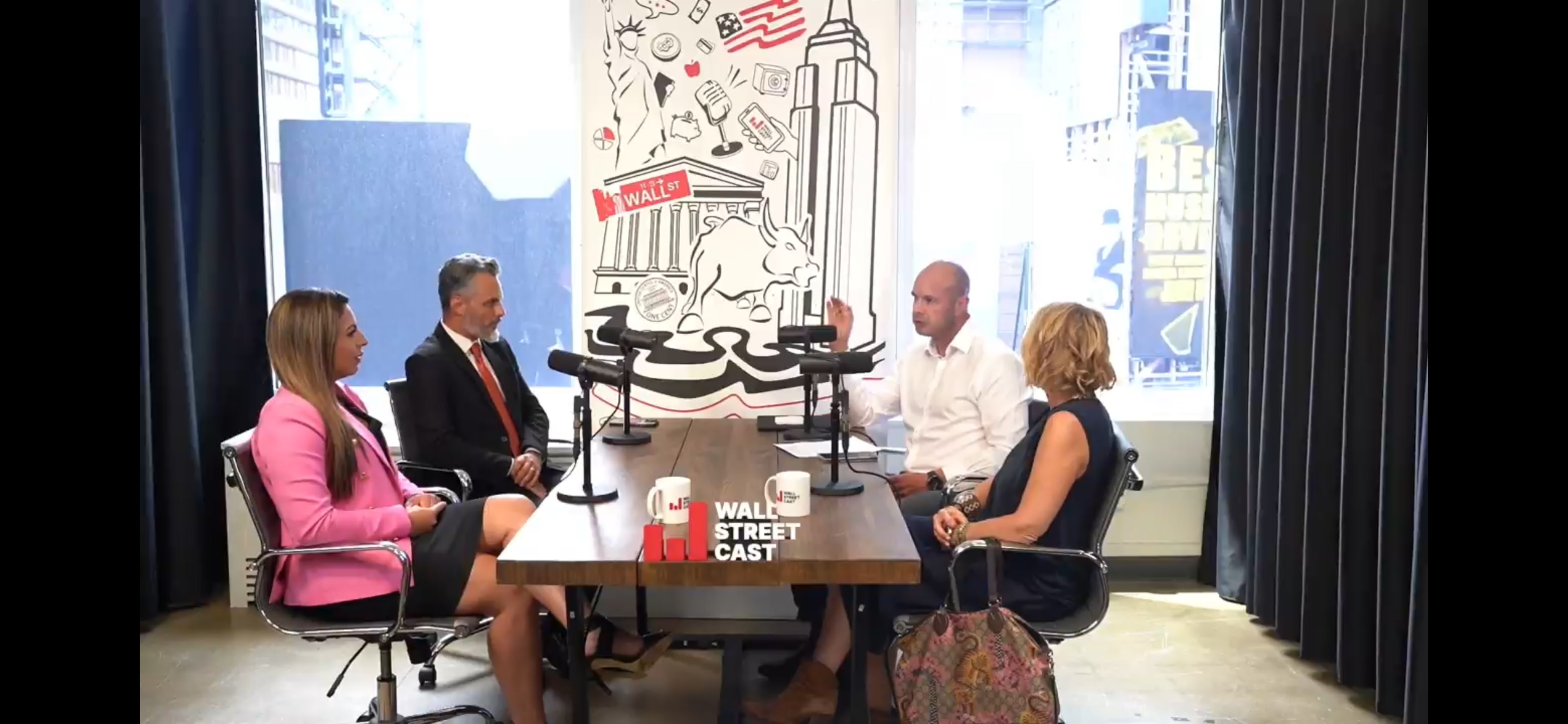 A group of four people is seated around a table with microphones in a modern office decorated with a mural of the Empire State Building and other cityscape elements.