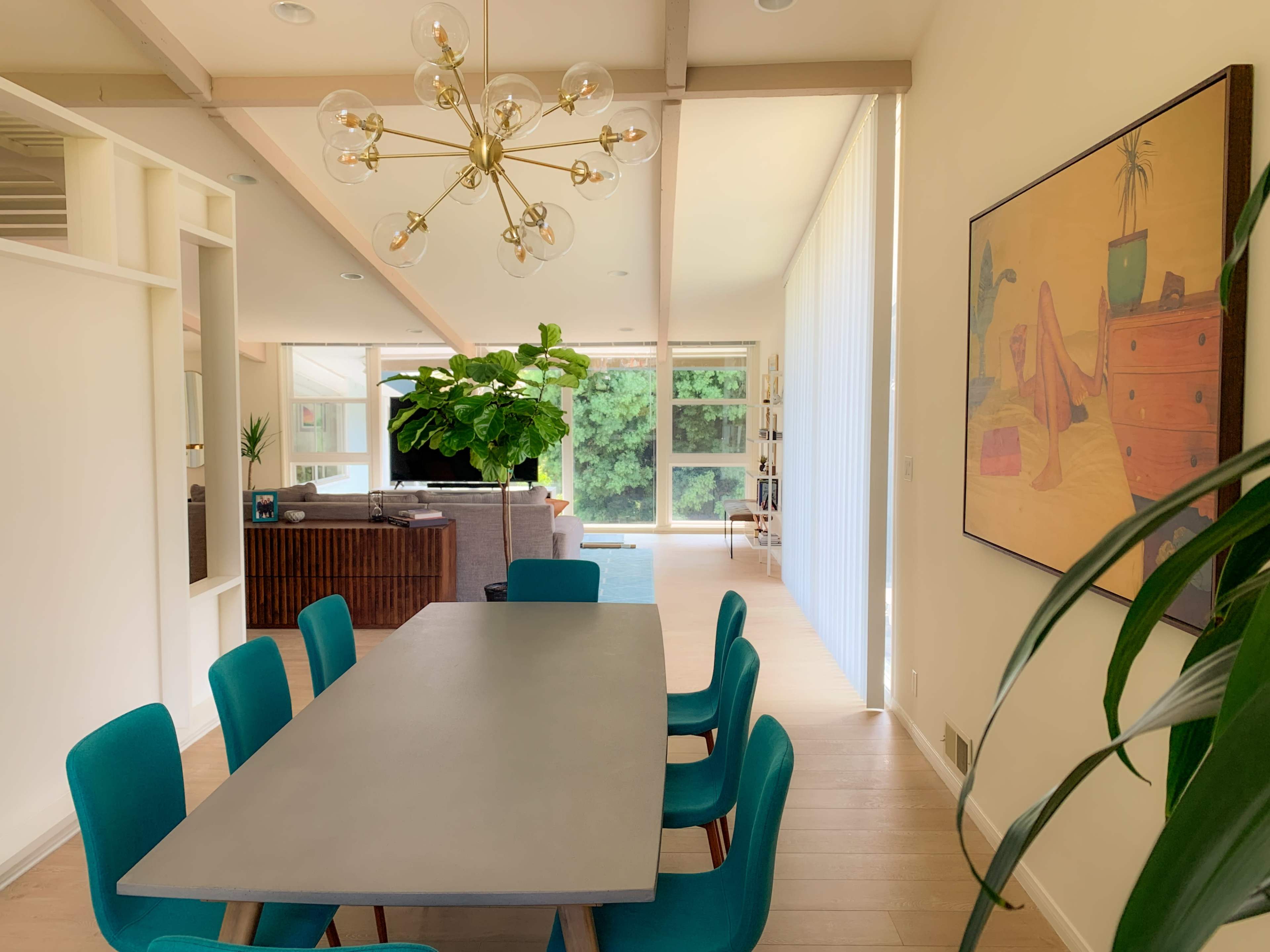 A modern dining area with a long gray table surrounded by teal chairs, leading into a living space with large windows and a prominent indoor plant.
