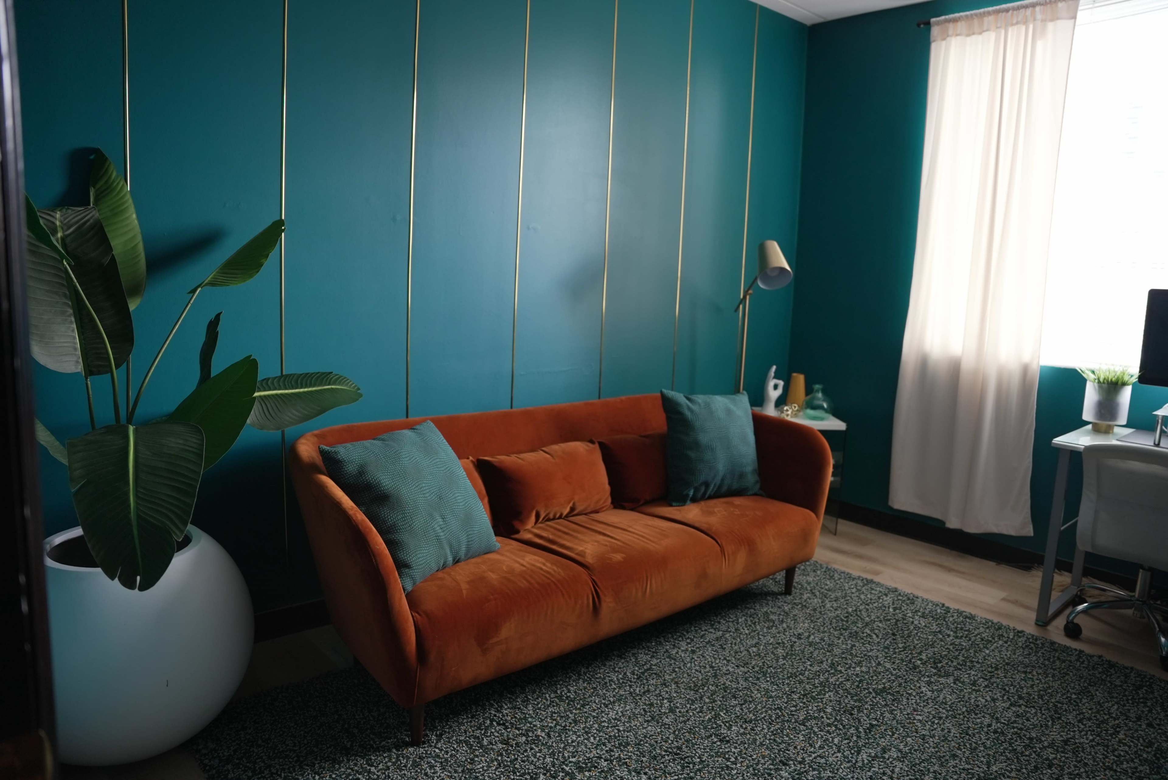 The image shows a room with a teal wall, an orange couch, a potted plant, and a light-colored rug.