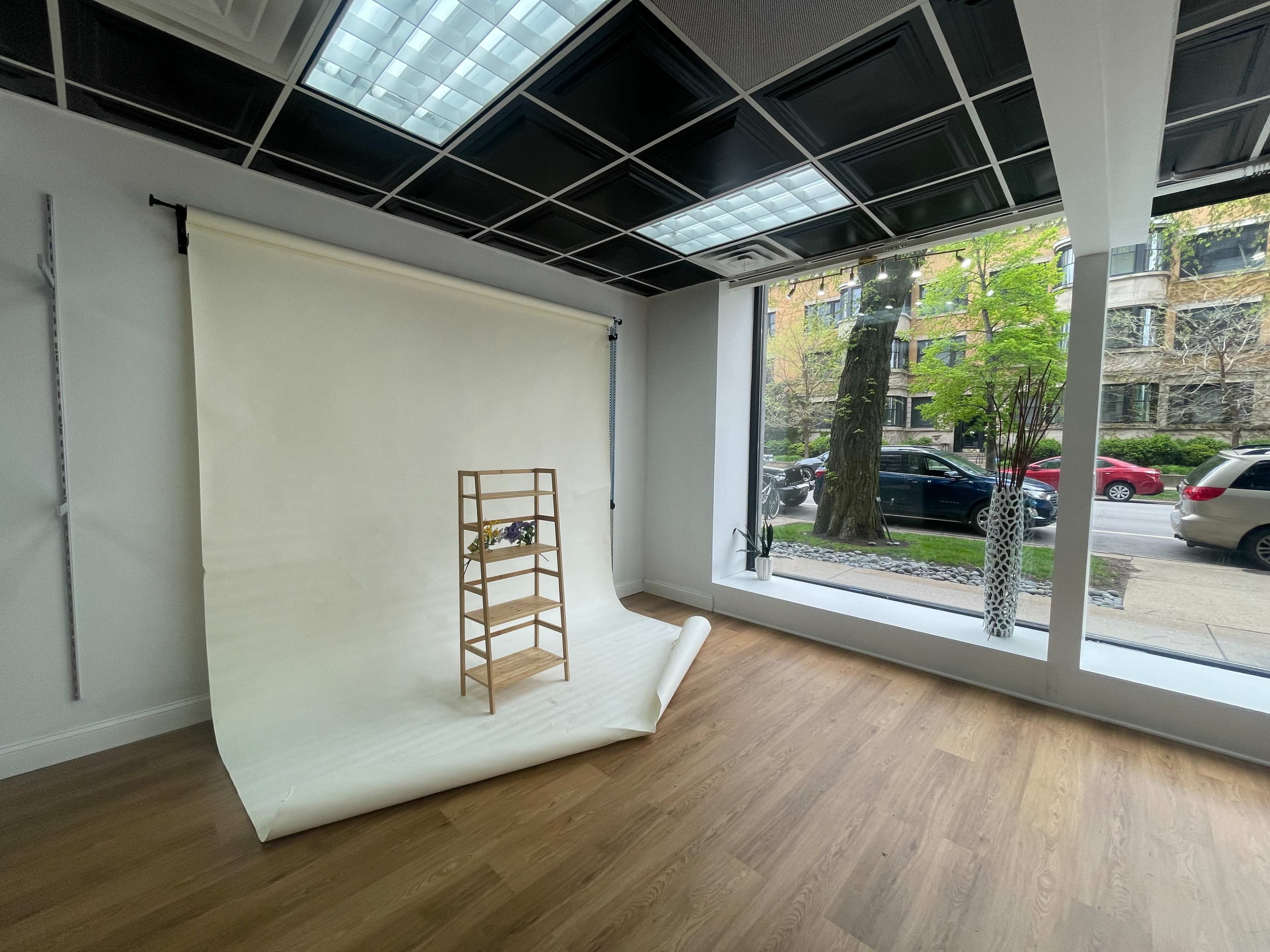 The image shows a minimalist interior with a light-colored backdrop, a wooden display stand, and large windows revealing a view of an urban street.