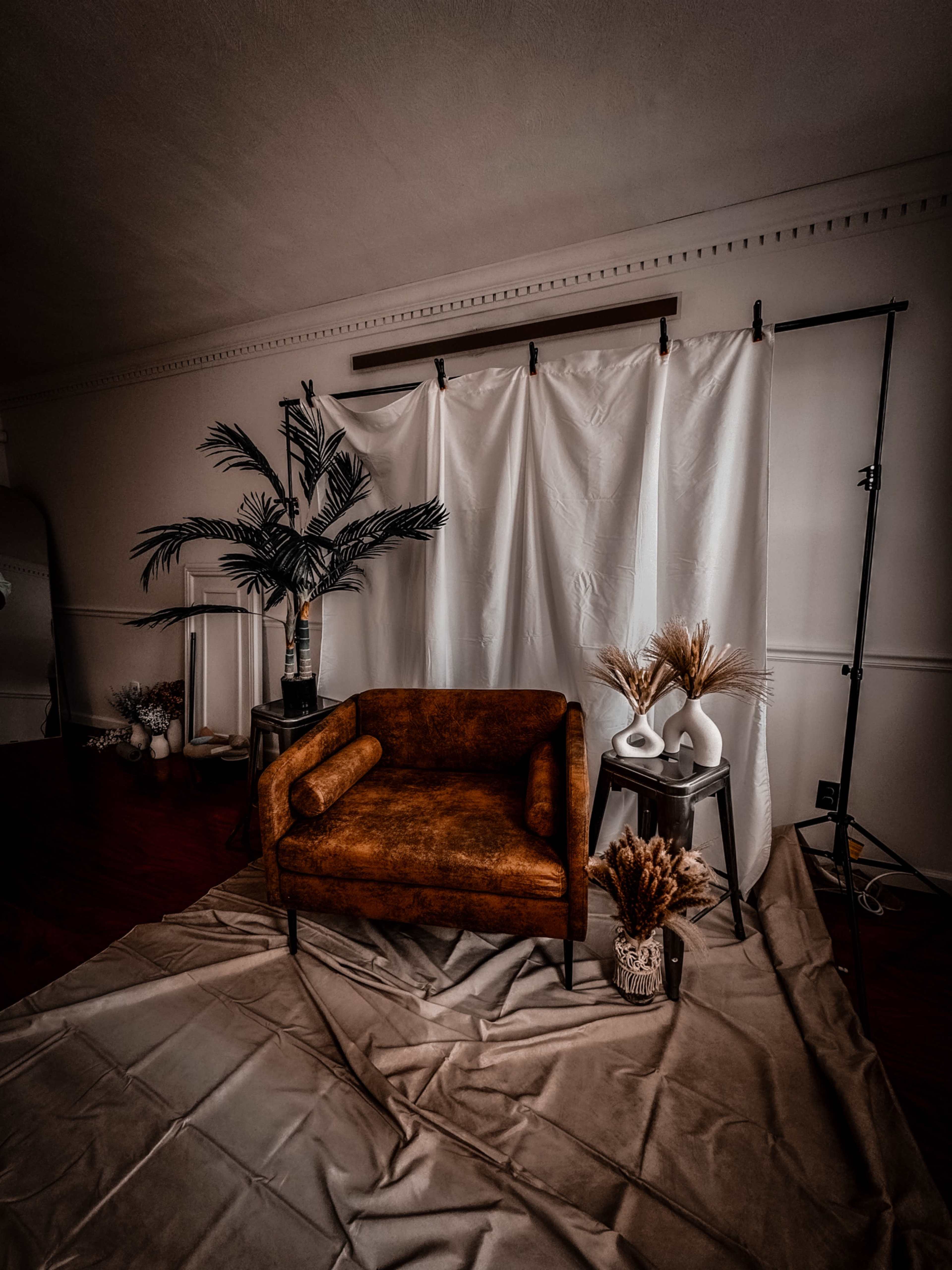 A brown upholstered chair is positioned in front of a white backdrop draped on a frame, surrounded by decorative plants and vases on a textured floor covering.