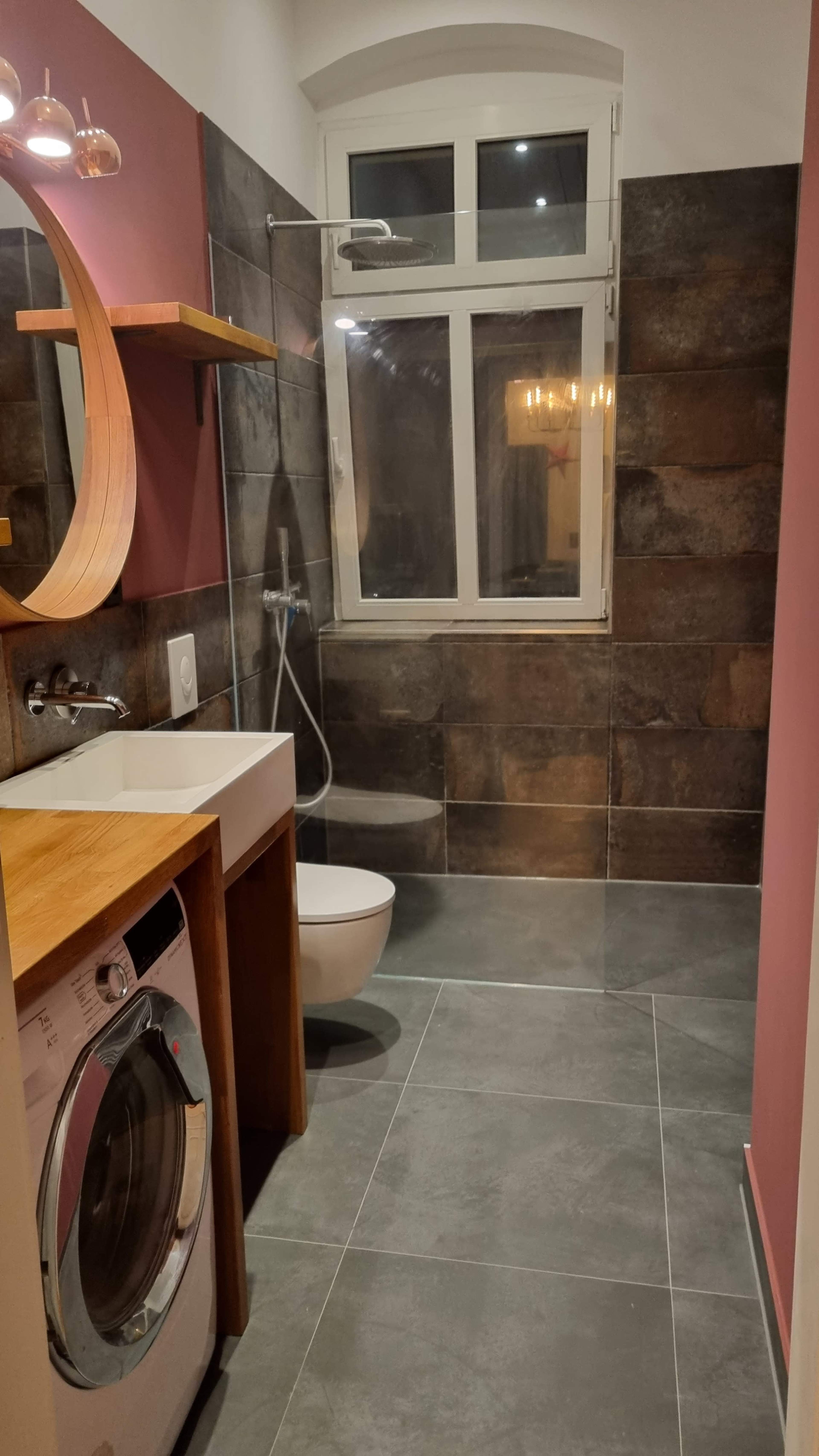 The image shows a modern bathroom with a wall-mounted sink, a washing machine, and a walk-in shower, featuring stone tiles and a circular mirror.
