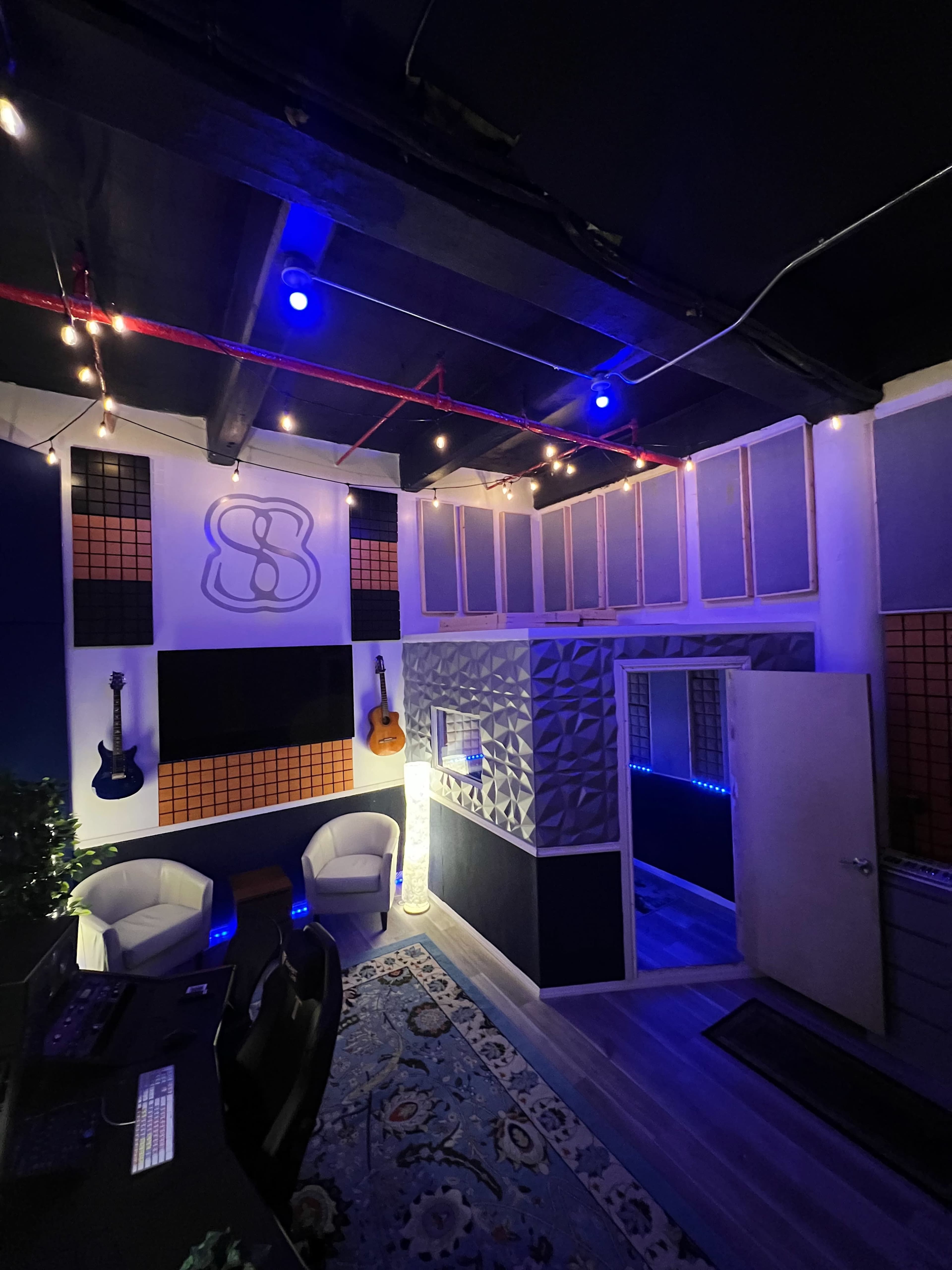 A music studio with wall-mounted guitars, sound panels, and a door leading to a separate room, illuminated by blue lights and decorative string lights.