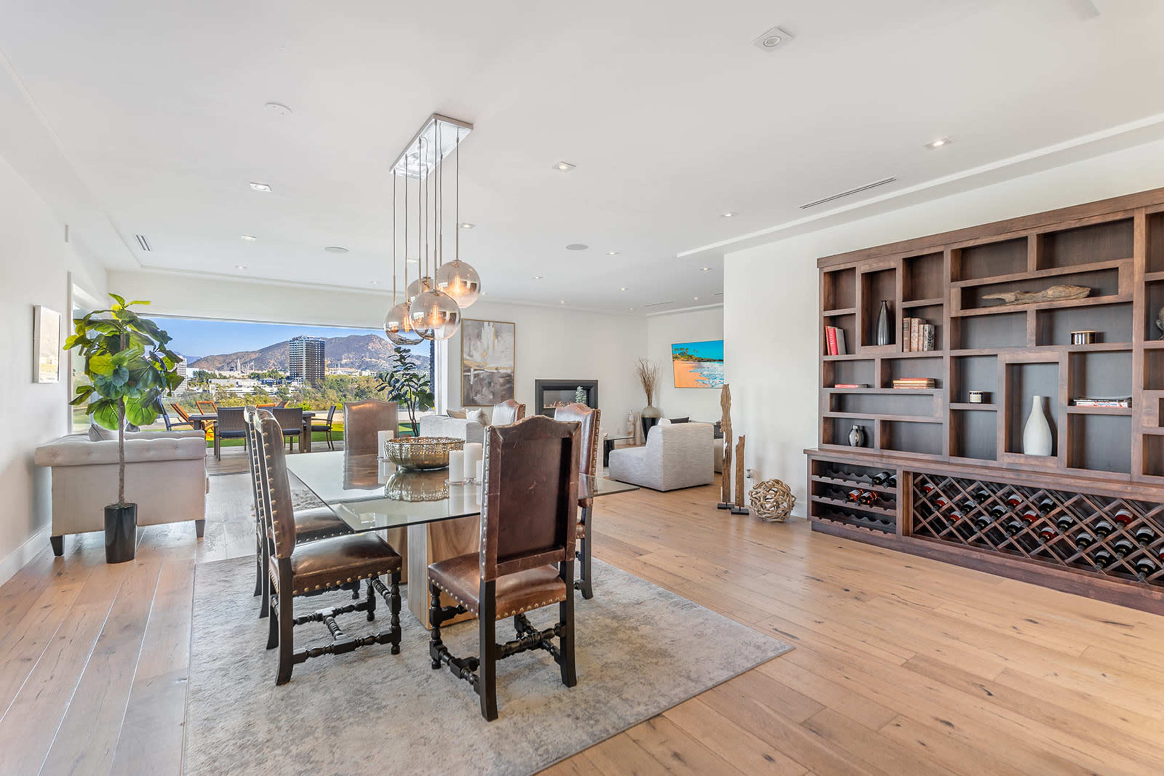 A modern dining area features a glass table surrounded by upholstered chairs, with a large window showcasing a mountain view and a wooden shelf displaying decorative items.
