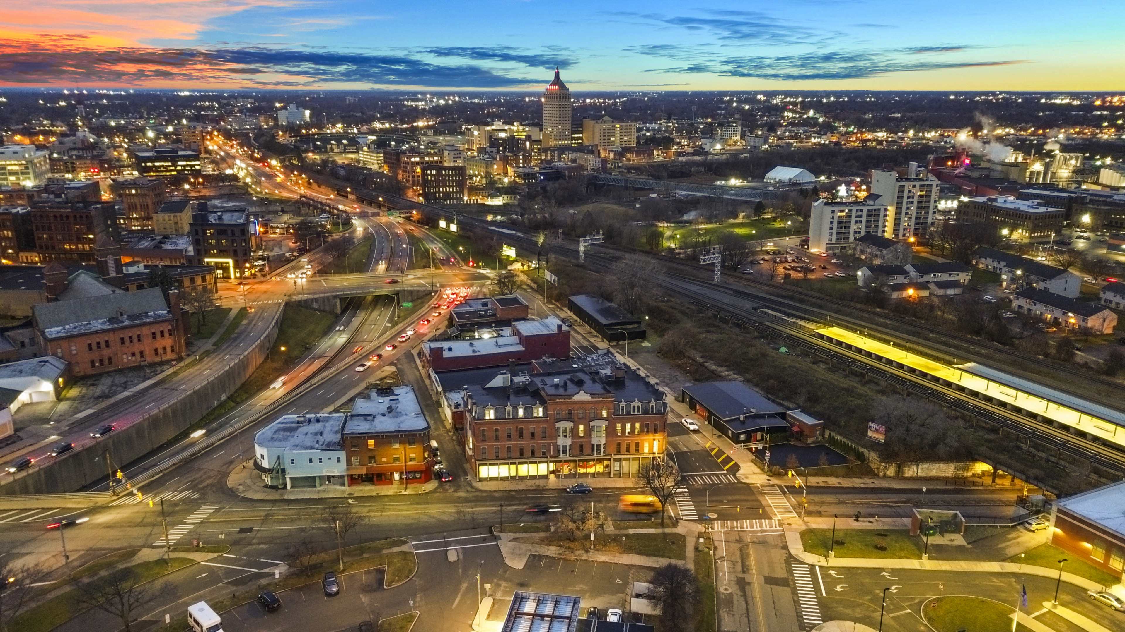 A cityscape at dusk, featuring a network of roads, buildings, and bright lights, with a river and railway visible in the foreground.